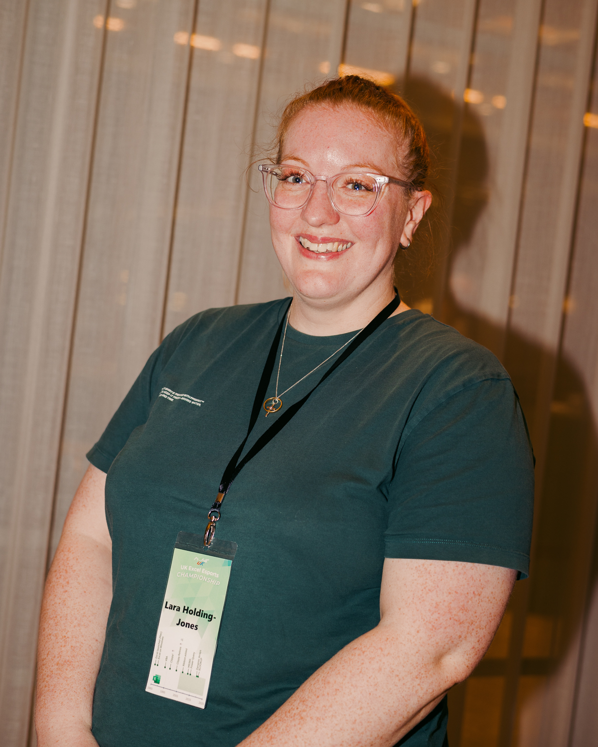 Woman with ginger hair and clear glasses wearing green medical scrubs and name badge, smiling against white panelled wall.