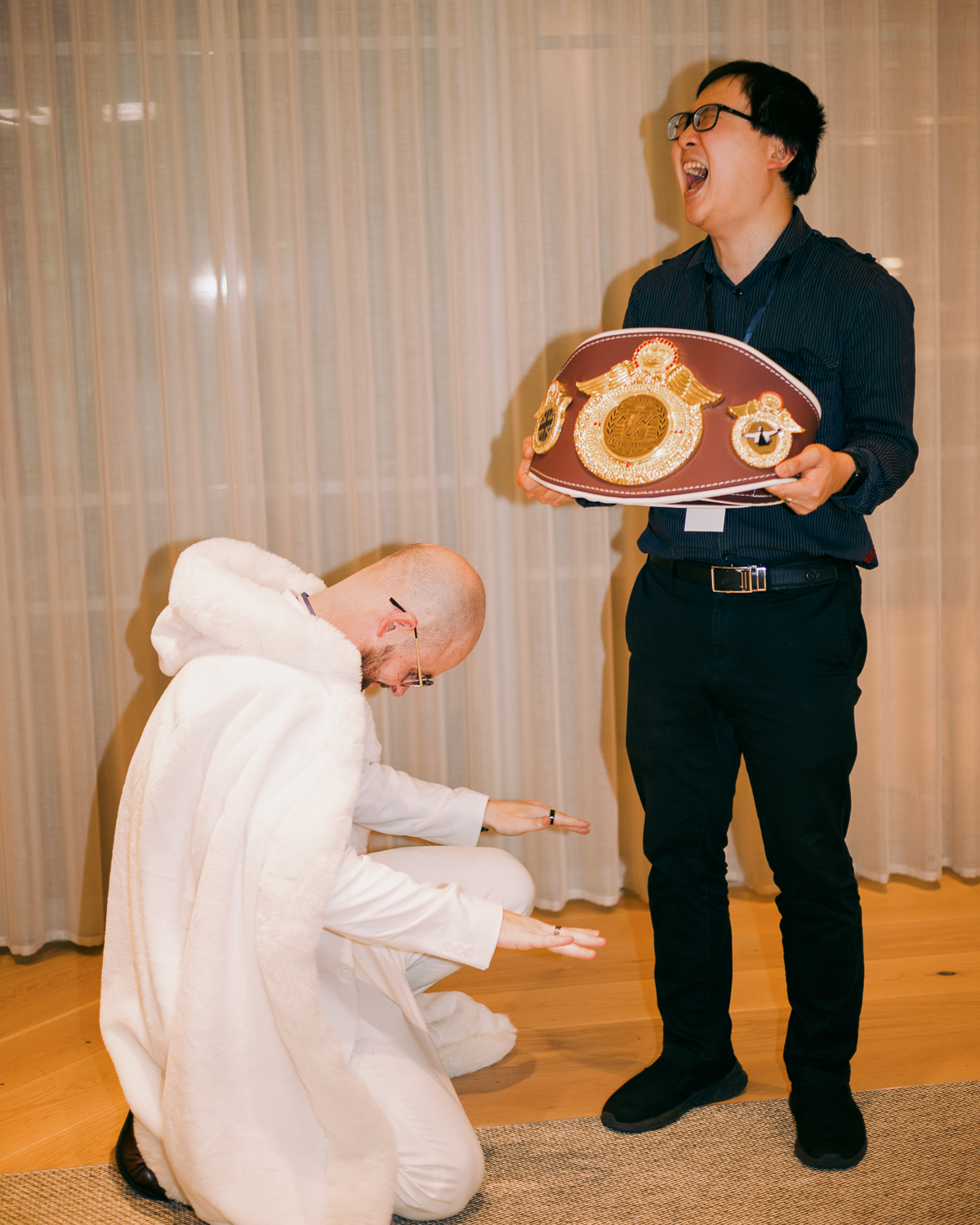 Person in black suit holding brown boxing belt whilst another person in white robe kneels beside them on wooden floor.