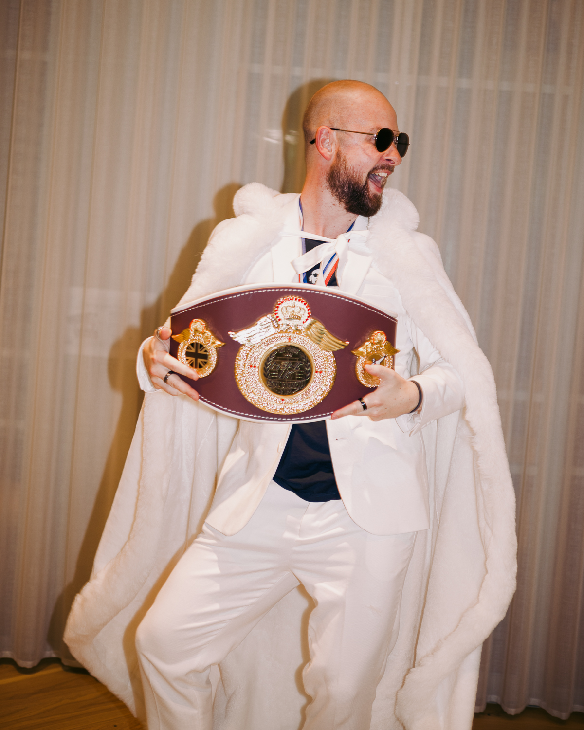 Bald man with sunglasses and beard wearing white hoodie holds ornate burgundy and gold boxing championship belt