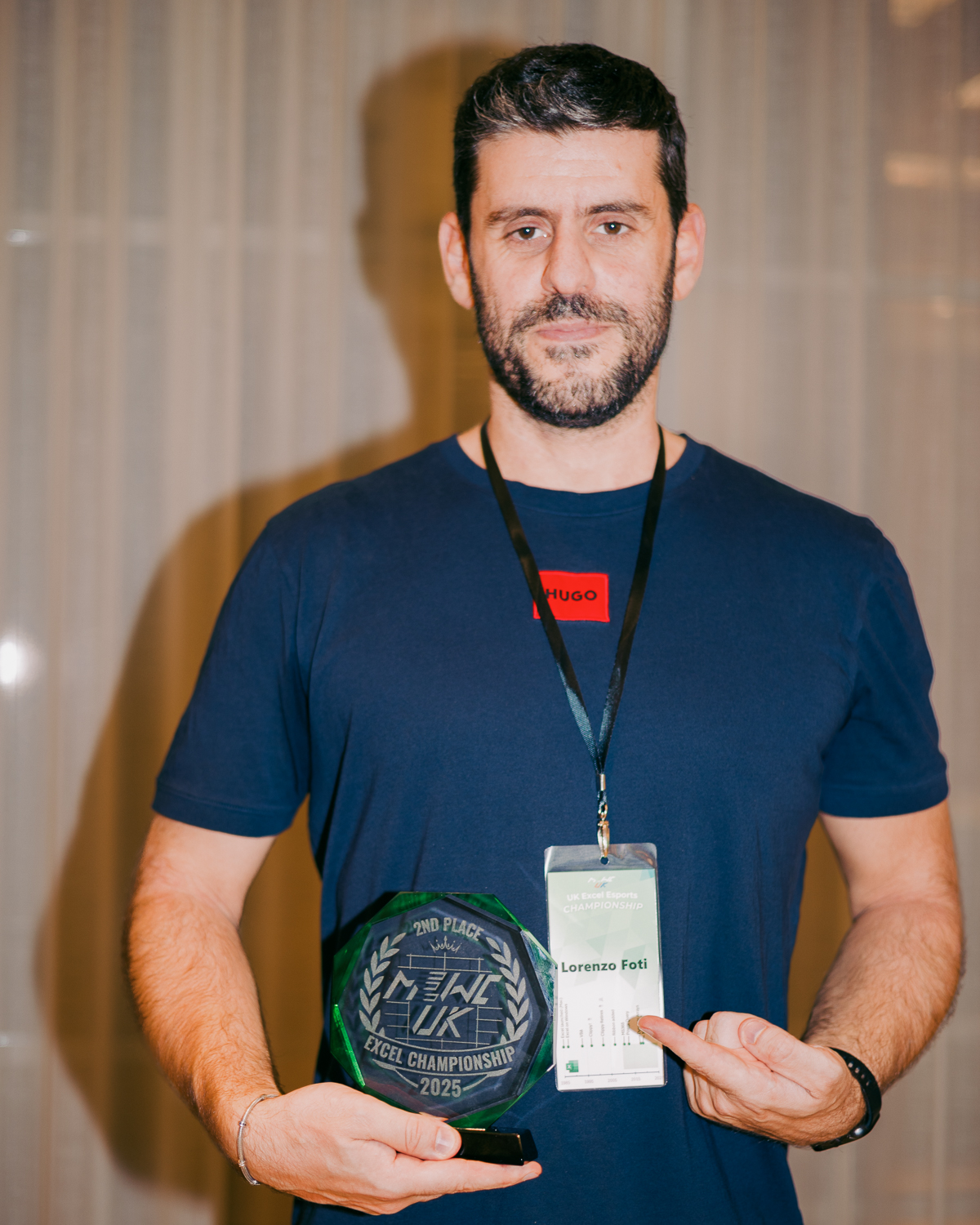 Man with beard in navy t-shirt holding green circular award trophy and white certificate, wearing red name badge and lanyard.
