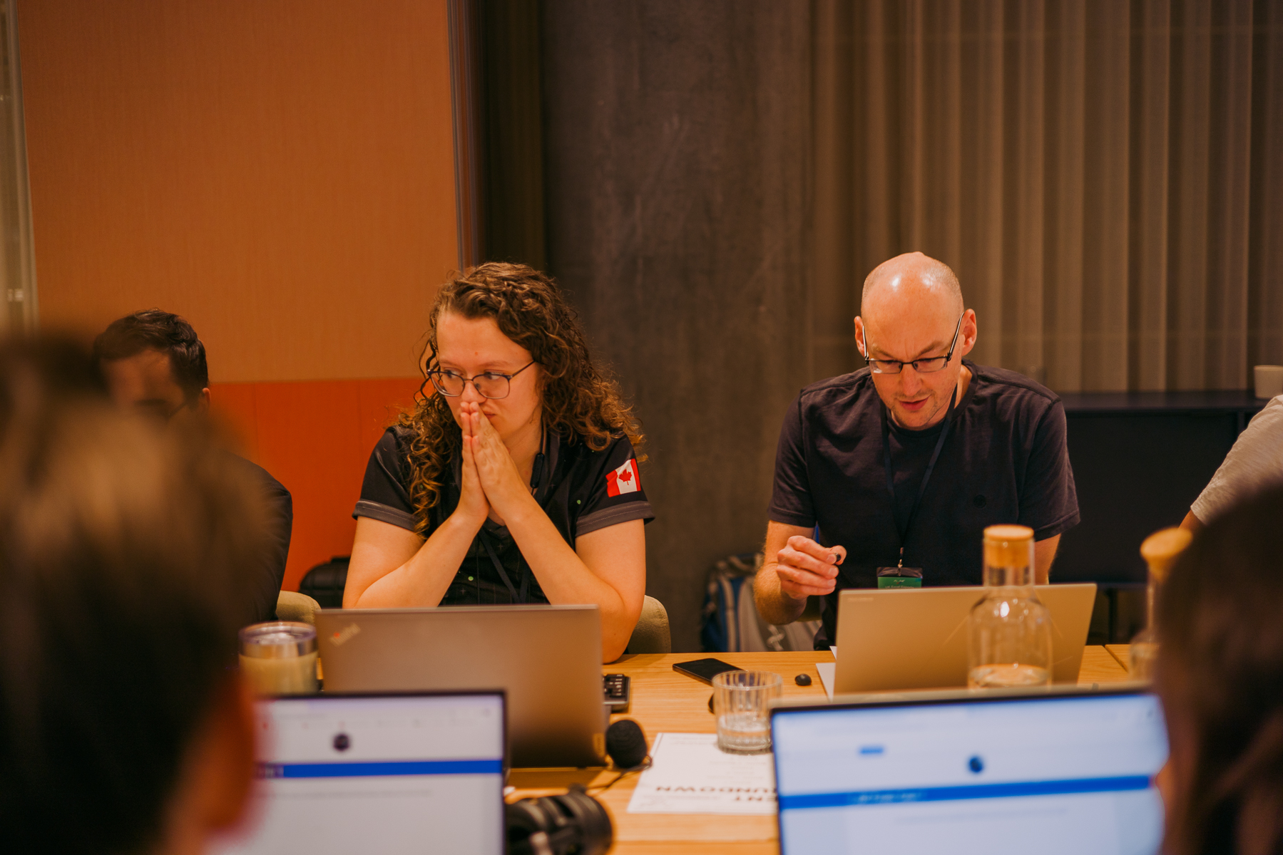 Woman with dark curly hair and man with glasses sit at table with laptops and drinks in meeting room with orange and brown walls.