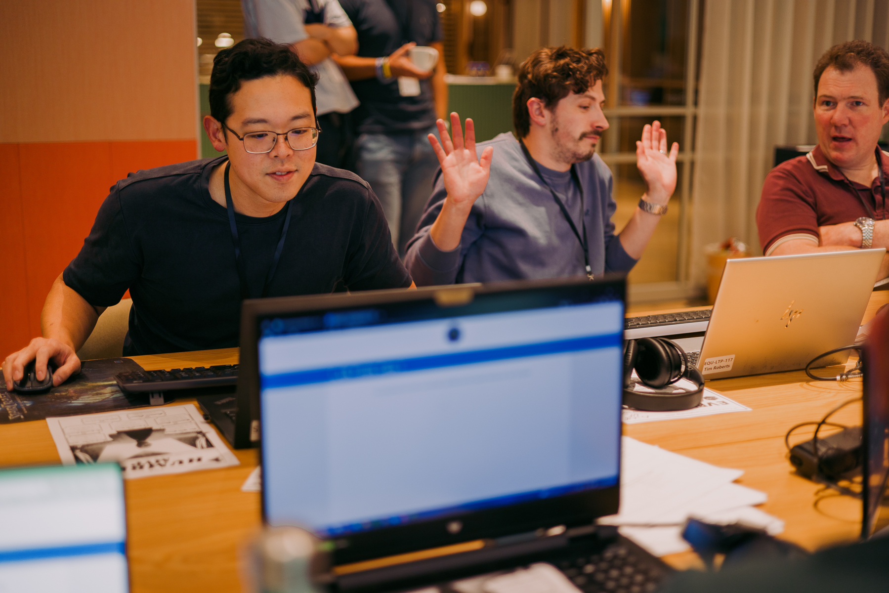 Three men in office setting with laptops, two raising hands. Warm lighting, wooden desk, papers scattered about.