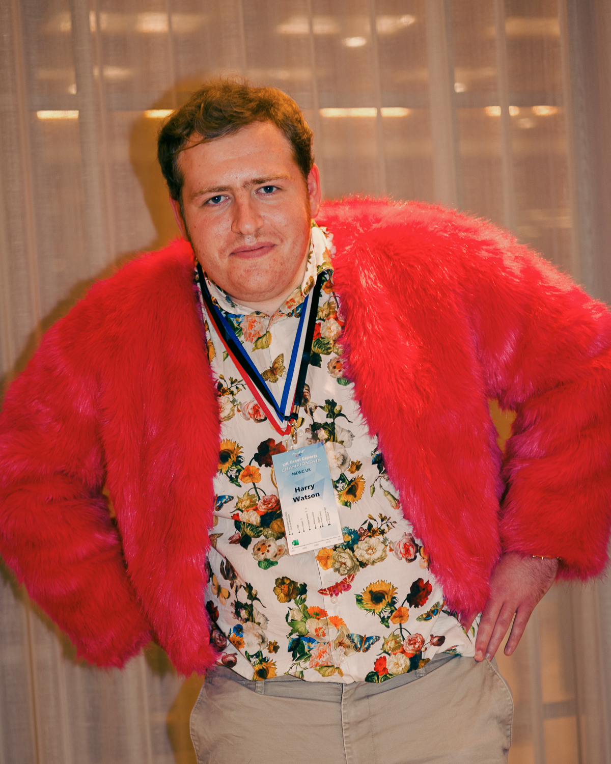 Man in bright red fuzzy coat and floral shirt with medal round neck, standing with hands on hips against blurred background.