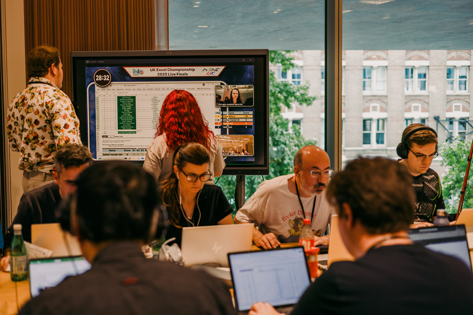 People working on laptops in office meeting room with large wall-mounted screen displaying website interface. Windows show buildings outside.