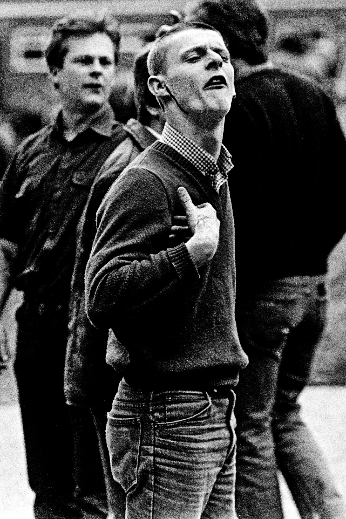 Black and white image of young men in 1980s clothing. Central figure wears jumper over checked shirt, head tilted back, mouth open.