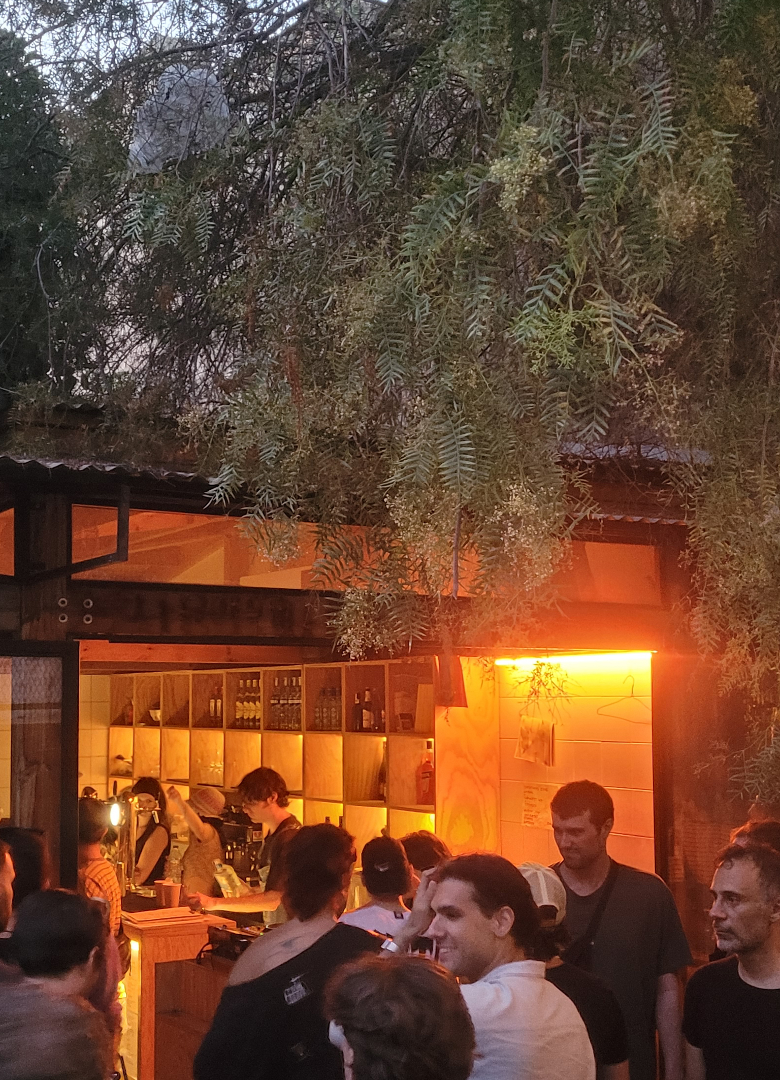 Outdoor bar with warm orange lighting surrounded by lush green foliage. People gathered around illuminated counter area under trees.