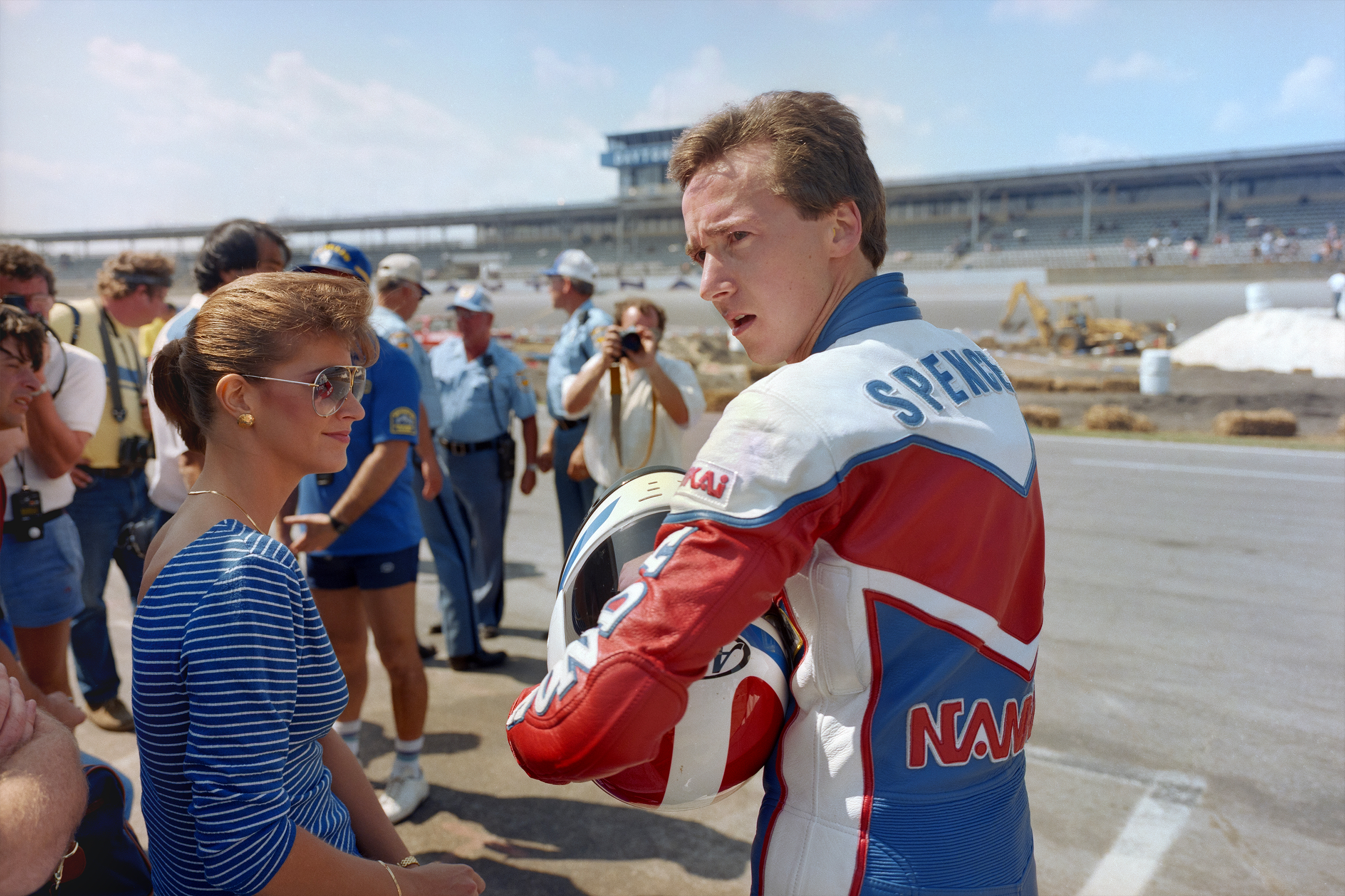 Racing driver in red and white suit talking to woman in blue striped shirt at sunny racetrack with grandstand and crowd behind.