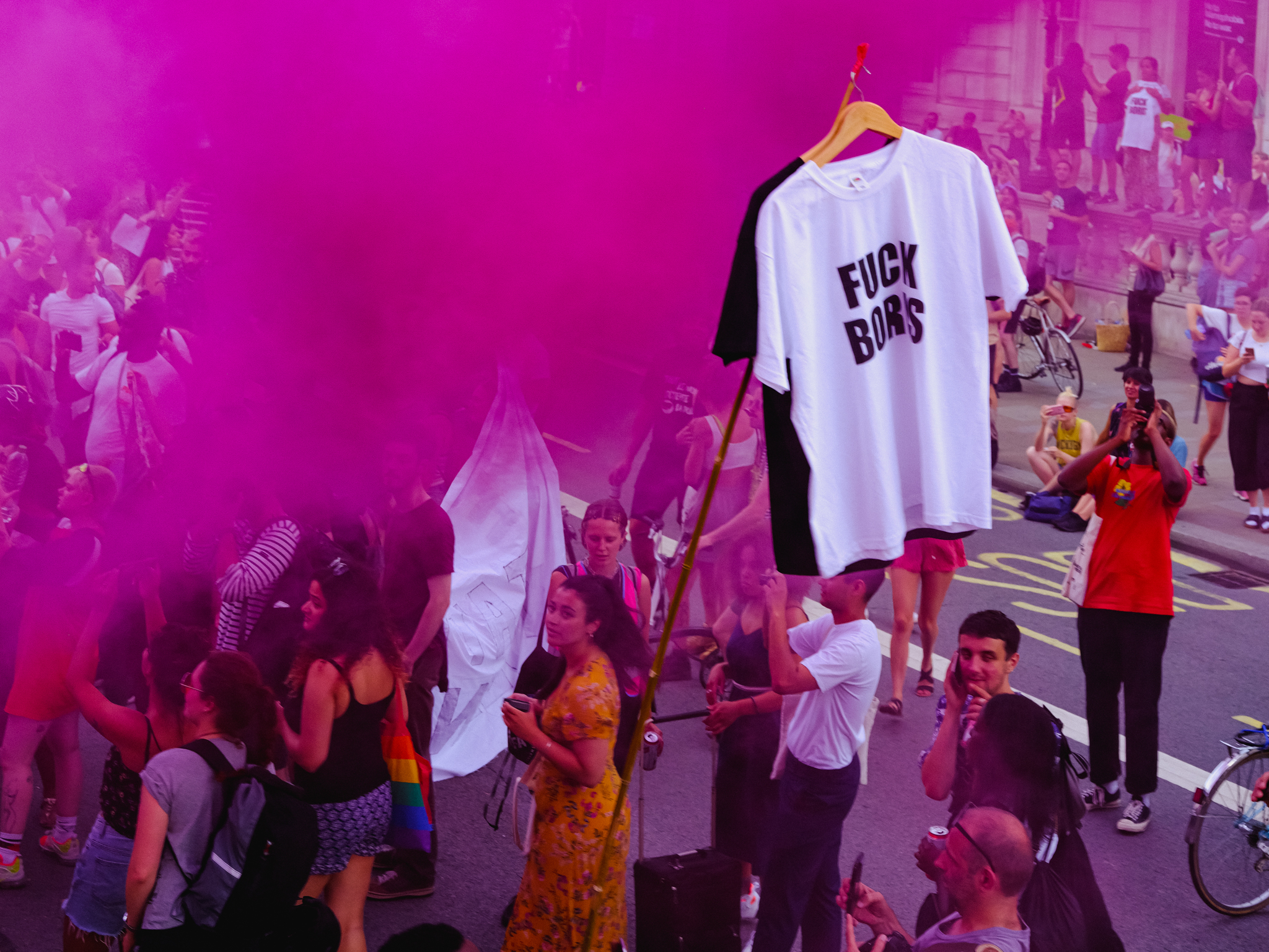 White t-shirt with "FUCK BORIS" text on hanger, overlaid on magenta-tinted crowd scene with people and bicycles on street.