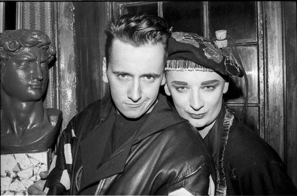 Black and white image of two people in dark clothing posing closely together indoors, with a decorative bust visible on the left.