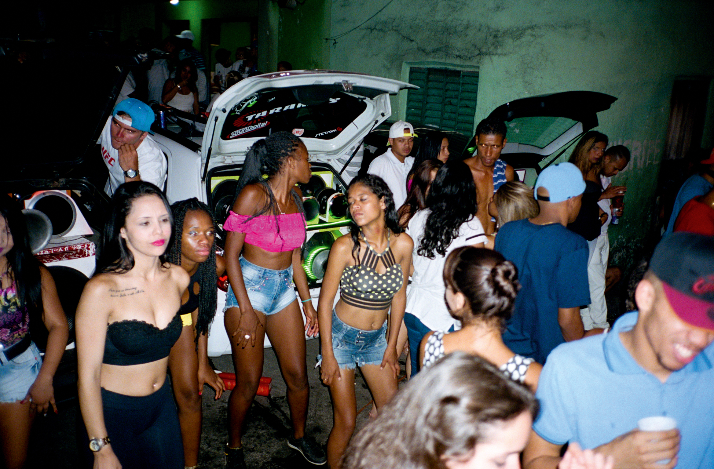 Crowded nighttime scene with young people gathered around white car with open bonnet, lit by green lighting against concrete wall.