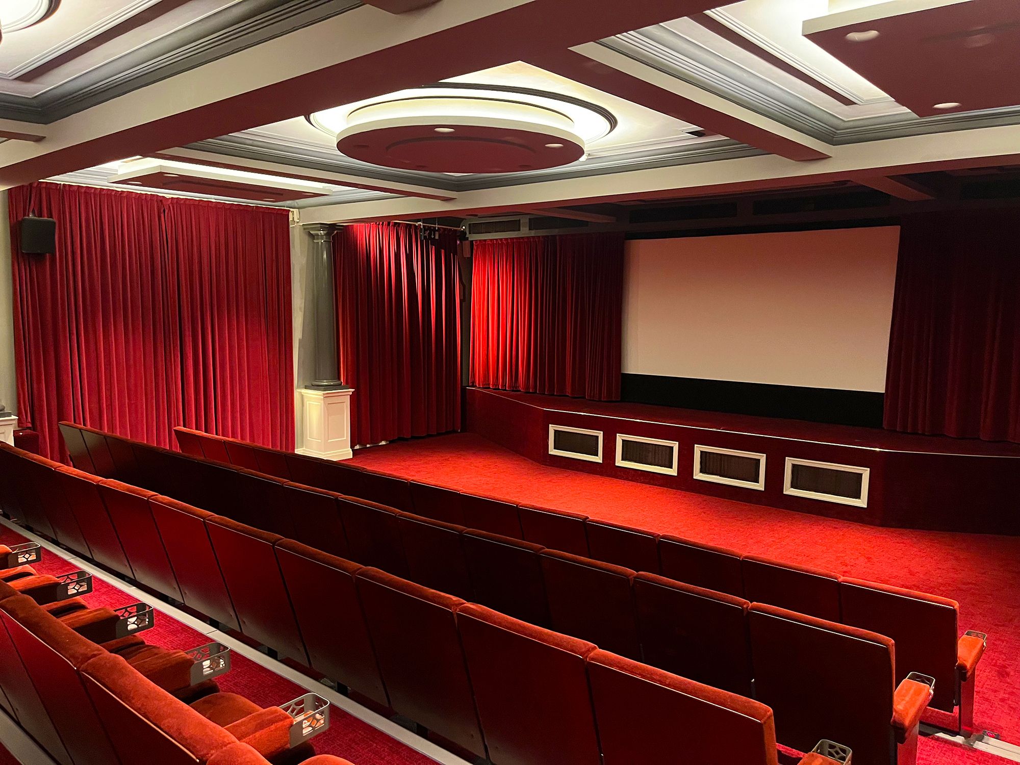 Opulent theatre auditorium with red velvet curtains, tiered seating, and a large projection screen.