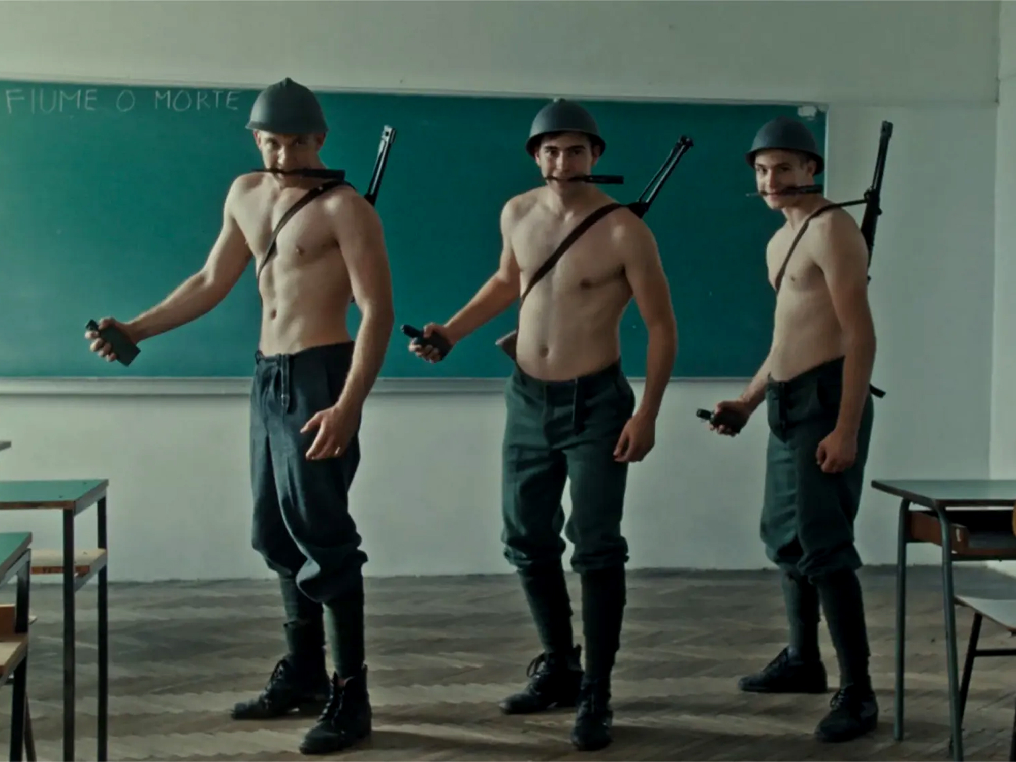 Three shirtless men in military caps and dark trousers holding hands in classroom, rifles strapped to backs, green chalkboard behind