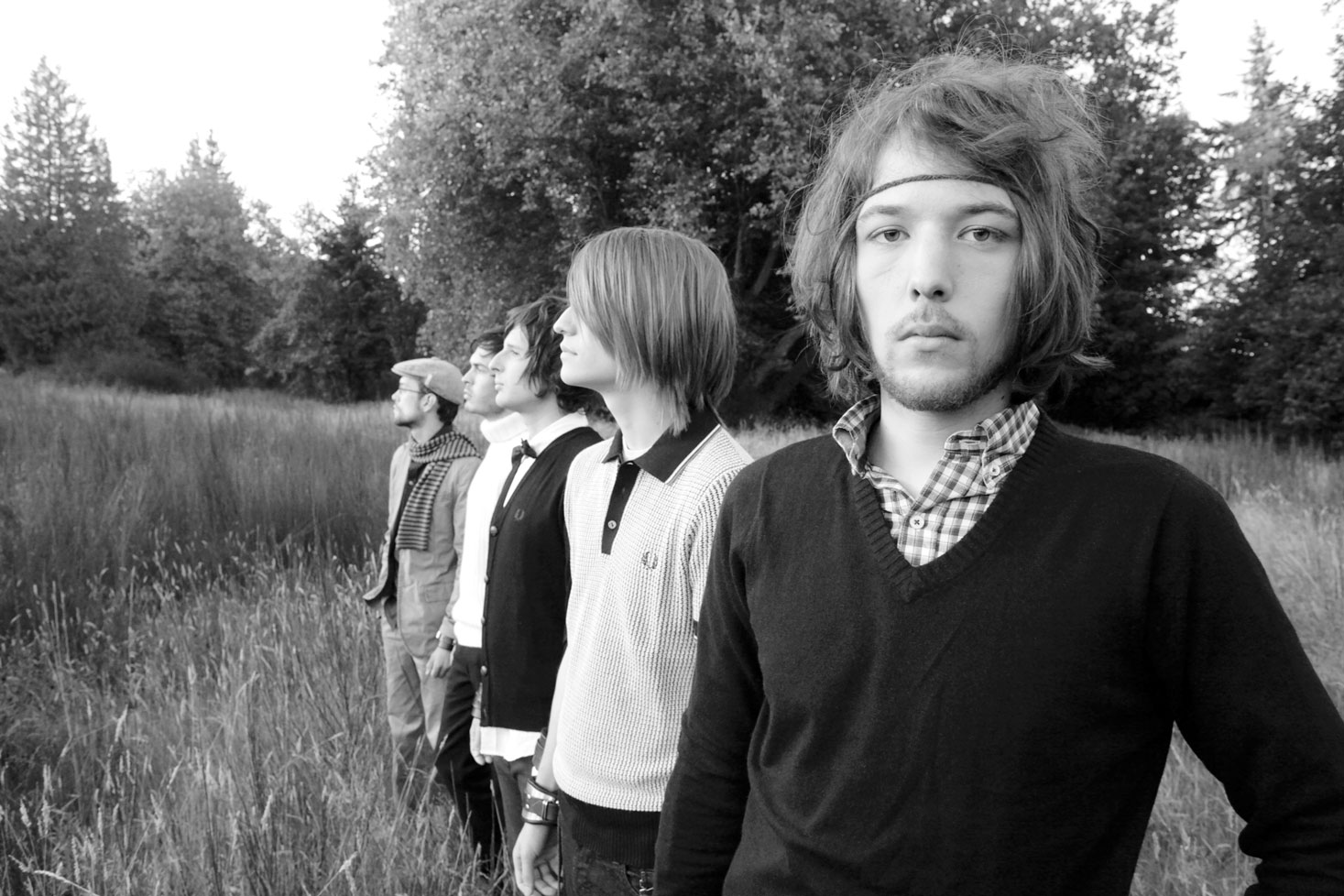 Four young men standing in grassy field with trees behind them. Black and white image. Man in foreground wears v-neck jumper.