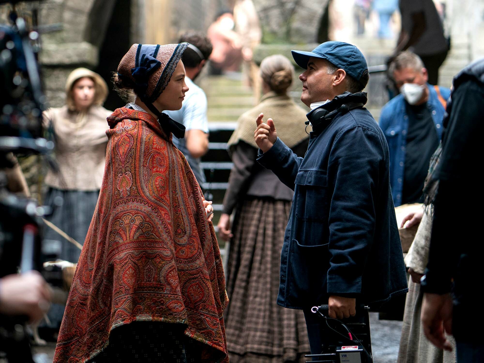 Two people in traditional clothing, one in a red and gold patterned robe, the other in a blue jacket, conversing on a crowded street.