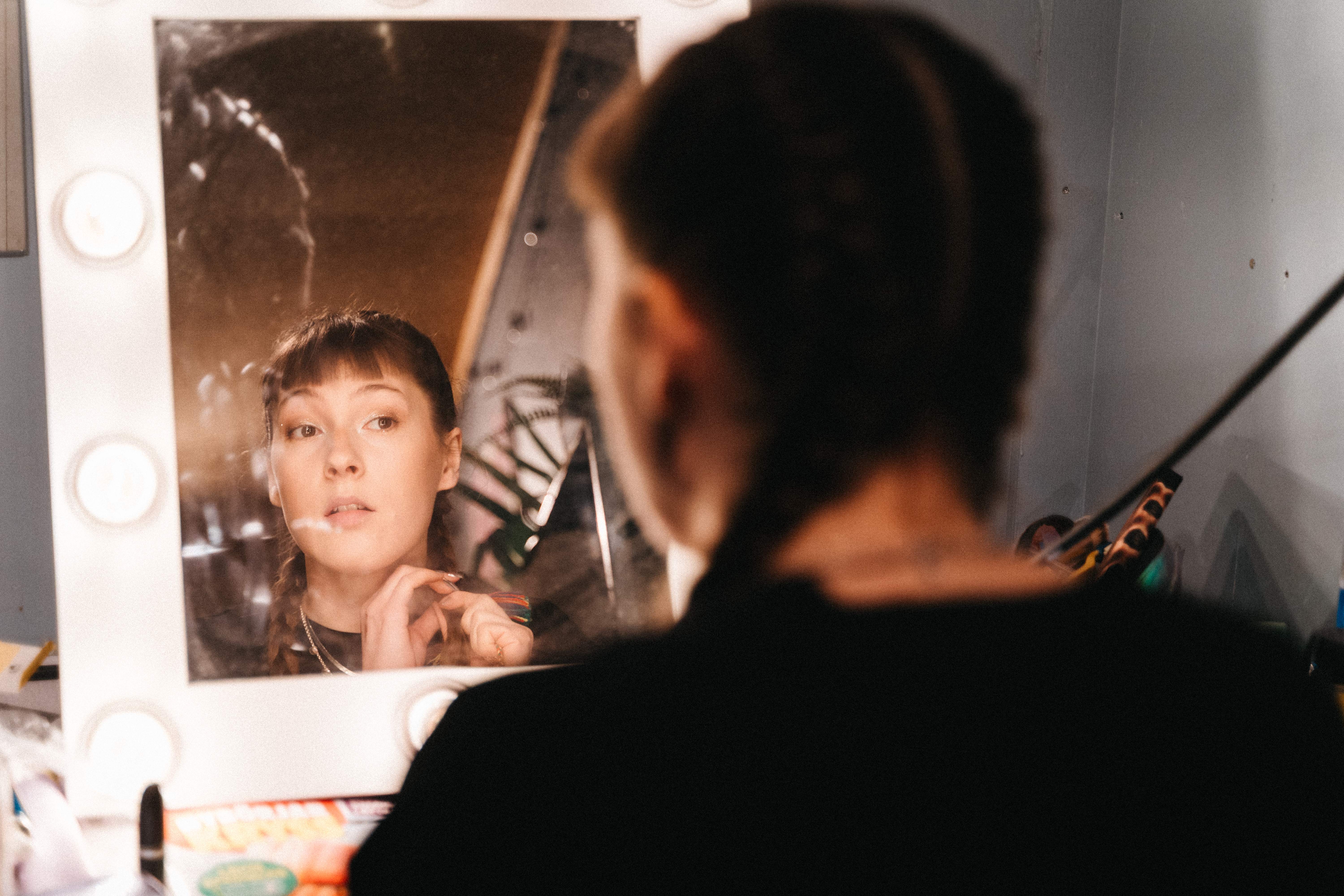 Person with dark hair looking at their reflection in mirror, applying makeup with brush in hand.