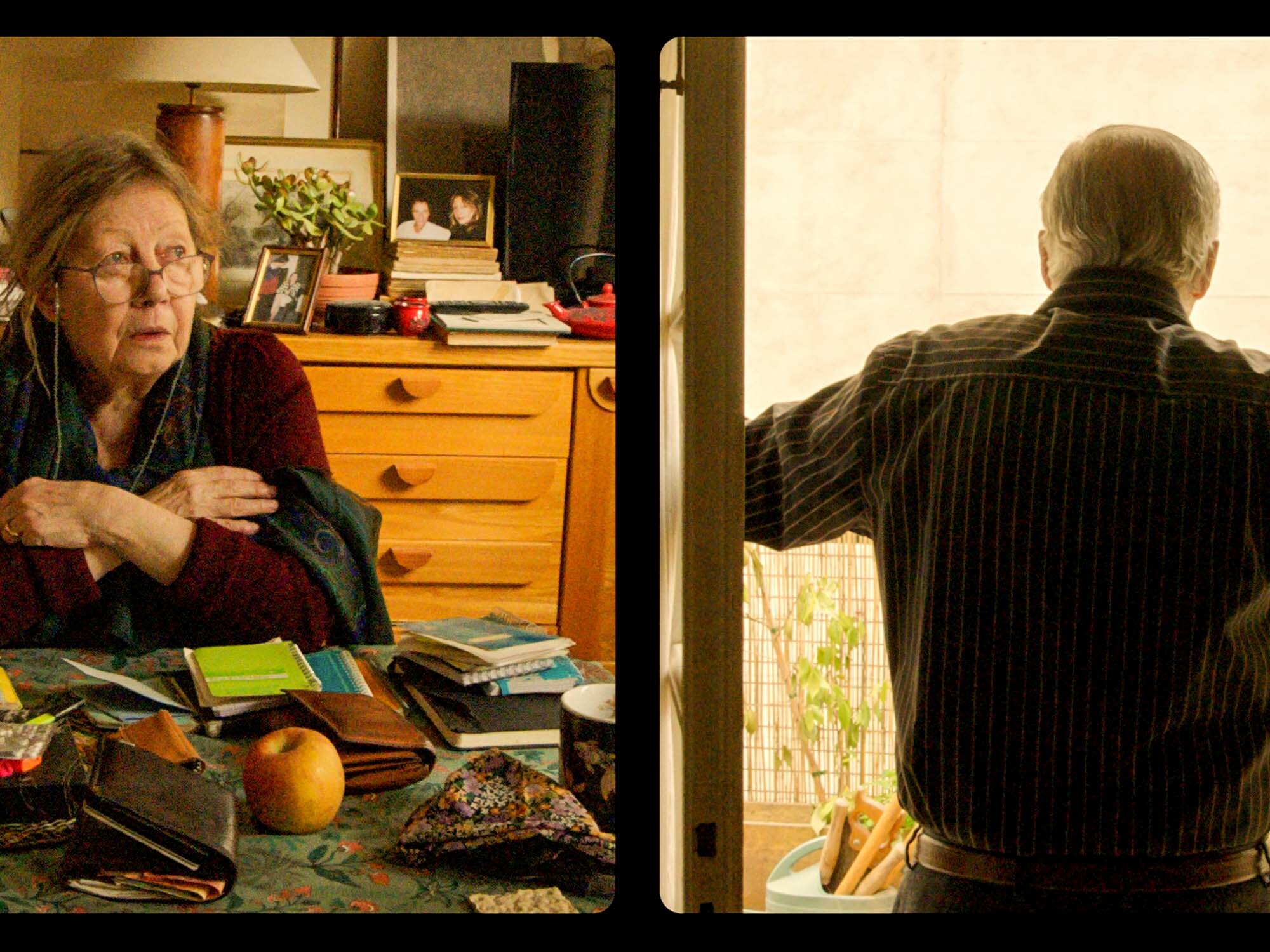 Elderly woman seated at a cluttered desk, looking pensive; man in a striped jacket stands at a window, gazing outside.