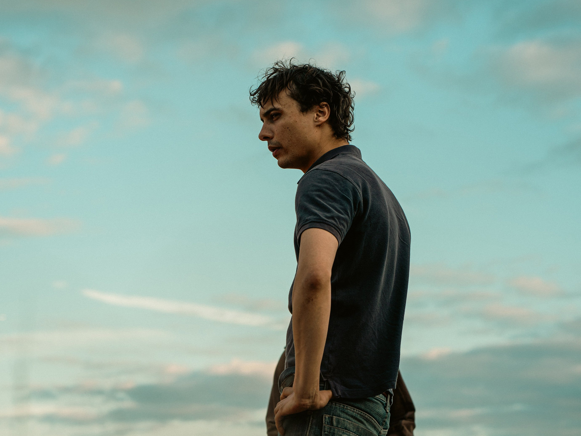 Man in dark t-shirt with curly hair looking to the side against turquoise sky with white clouds.