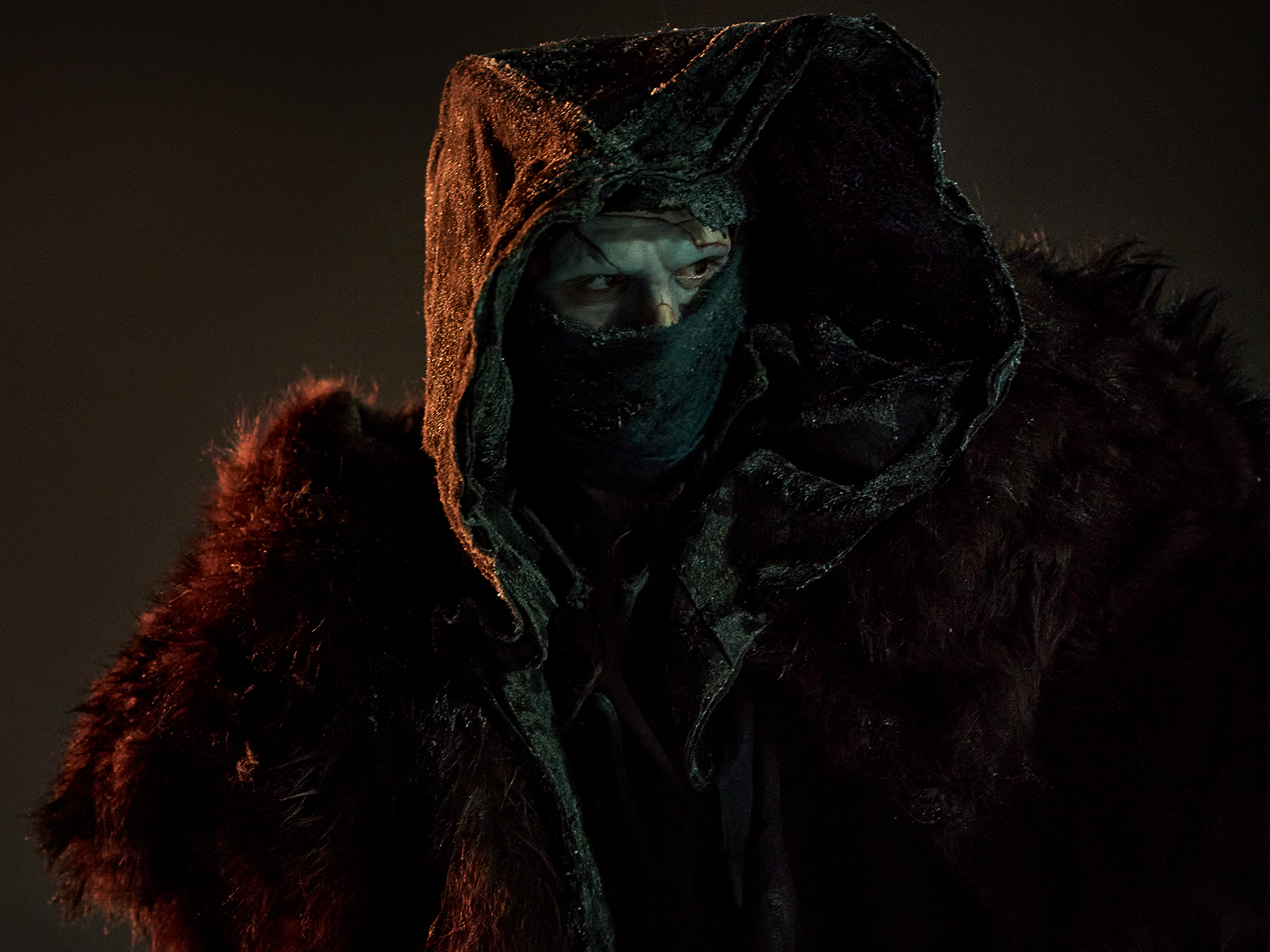 Figure in dark hooded robe with green-tinted face mask, orange fabric accent on hood, burgundy fur trim, against black background.