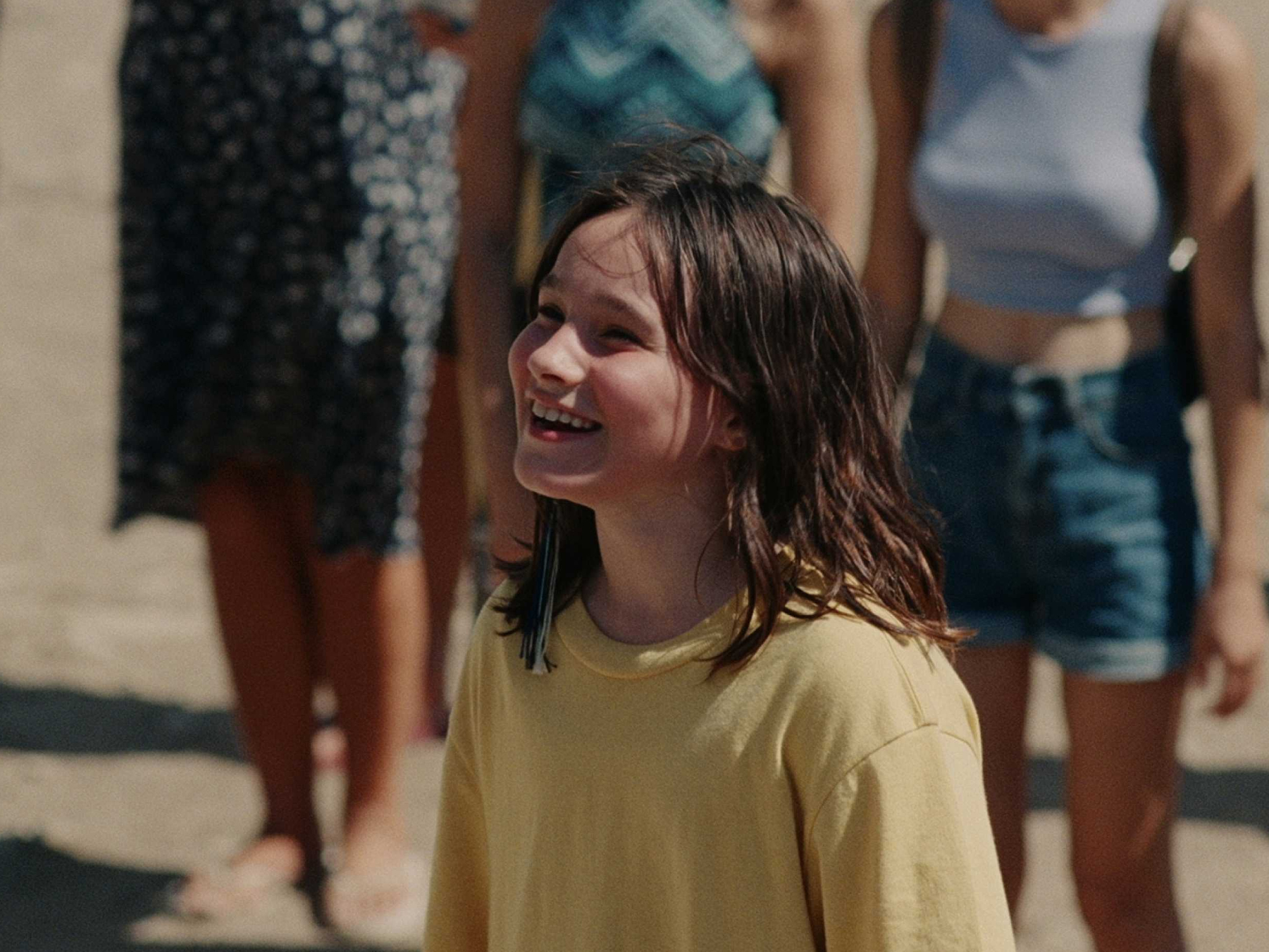 Frankie Corio, a young girl with brown hair, stands at the centre of the frame in a yellow t-shirt surrounded by people.