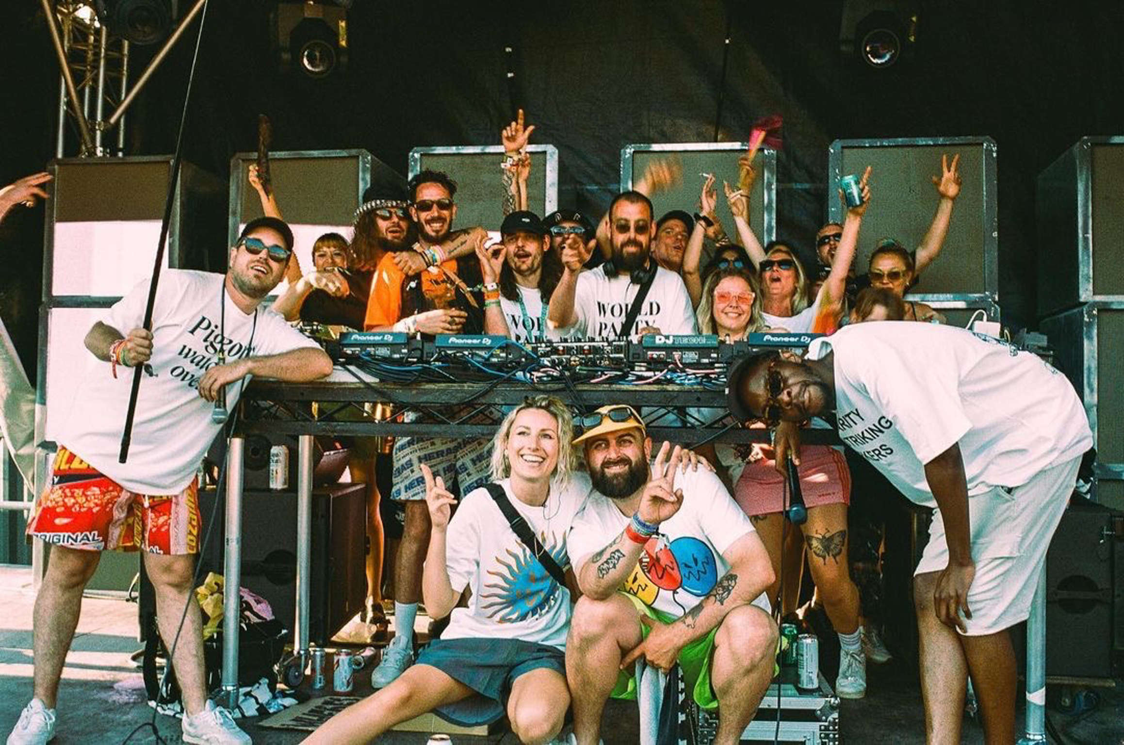 Large group of people on boat deck posing with raised arms and peace signs, wearing casual summer clothes and sunglasses.