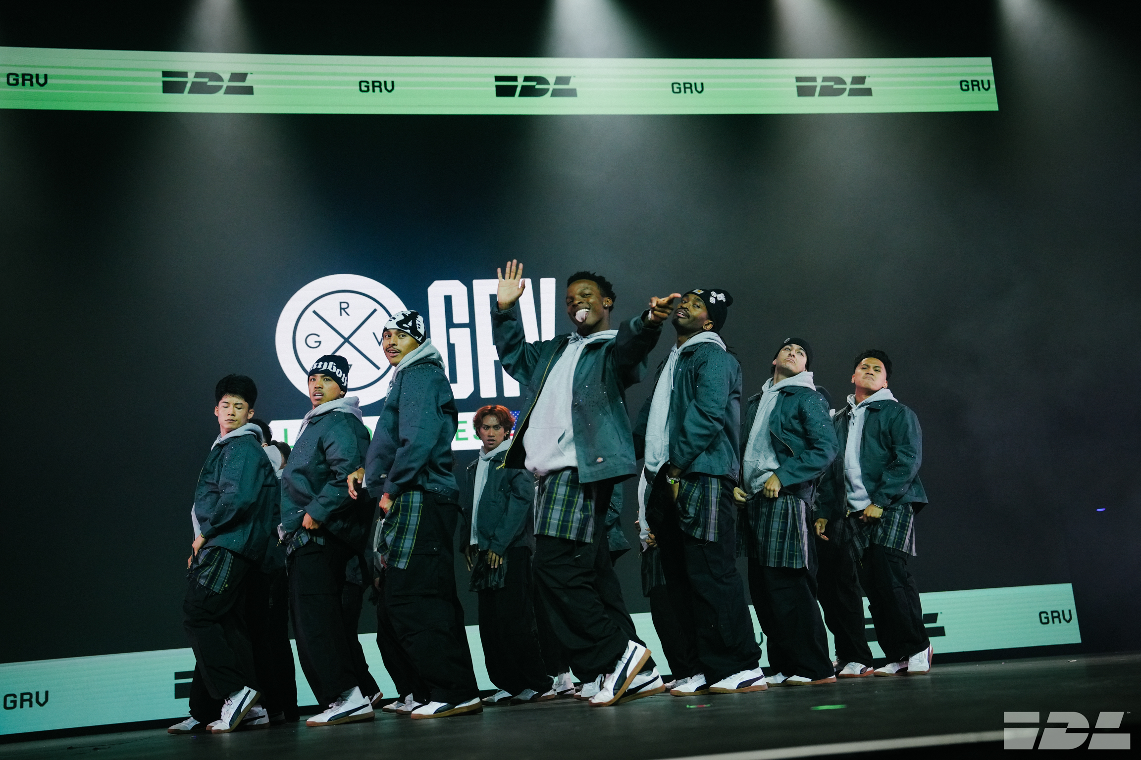 Nine performers in matching dark green tracksuits on stage with green lighting and "GRU" branding visible in background.