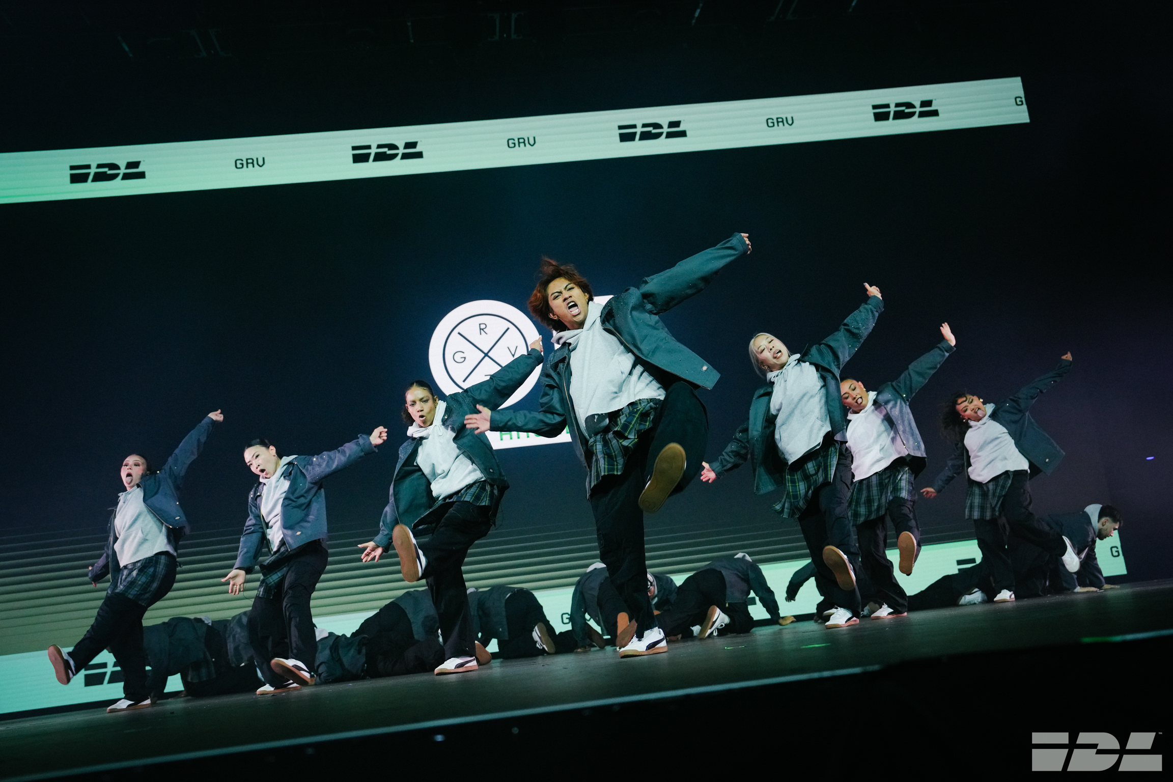 Seven performers in white shirts and dark trousers dancing in synchronised formation on stage with circular logo backdrop and blue lighting.