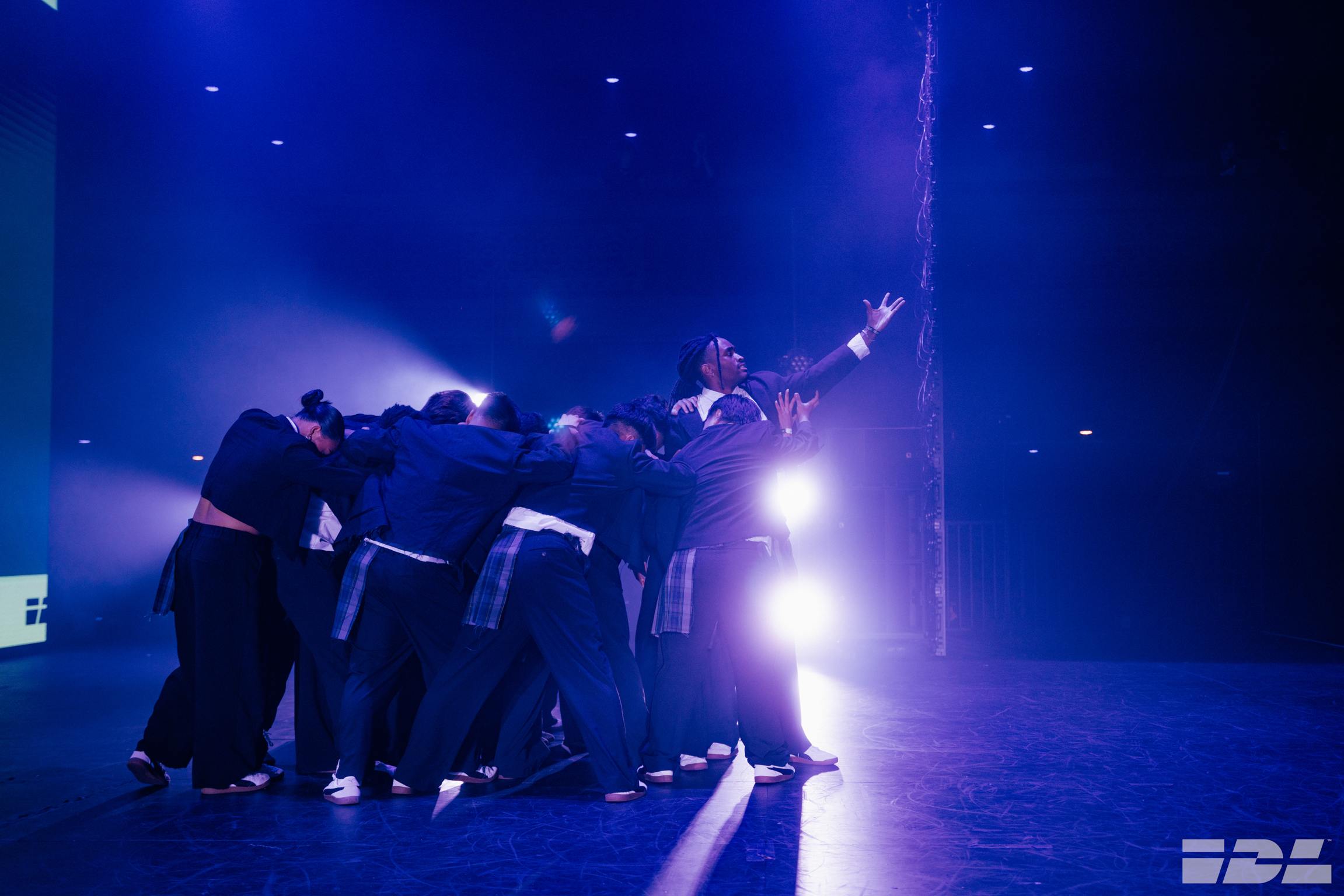 Group of performers in dark clothing huddled together on stage, illuminated by bright white spotlight against deep blue lighting.