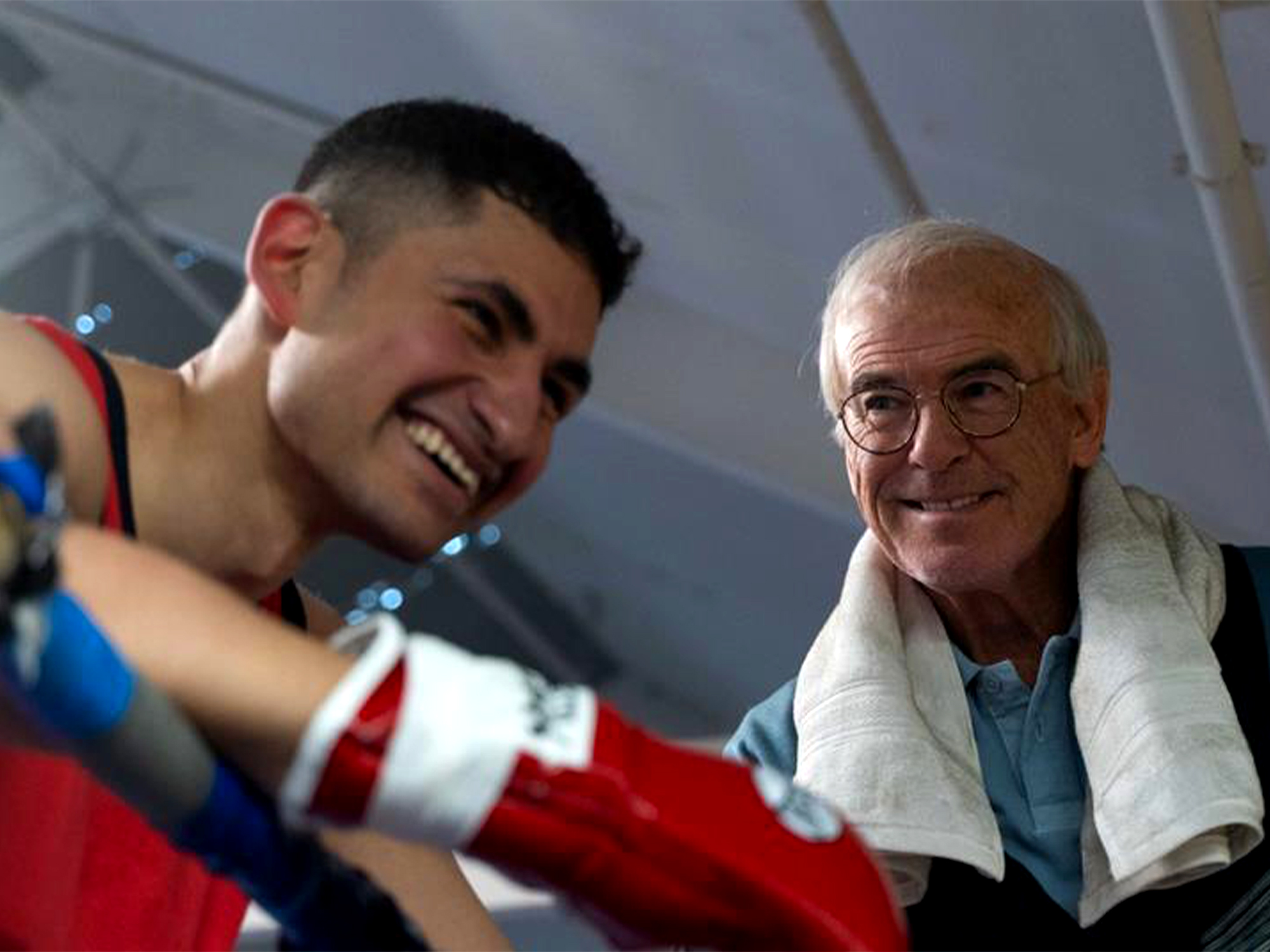Young boxer in red gloves with short dark hair smiling alongside elderly man with glasses and white towel around neck.