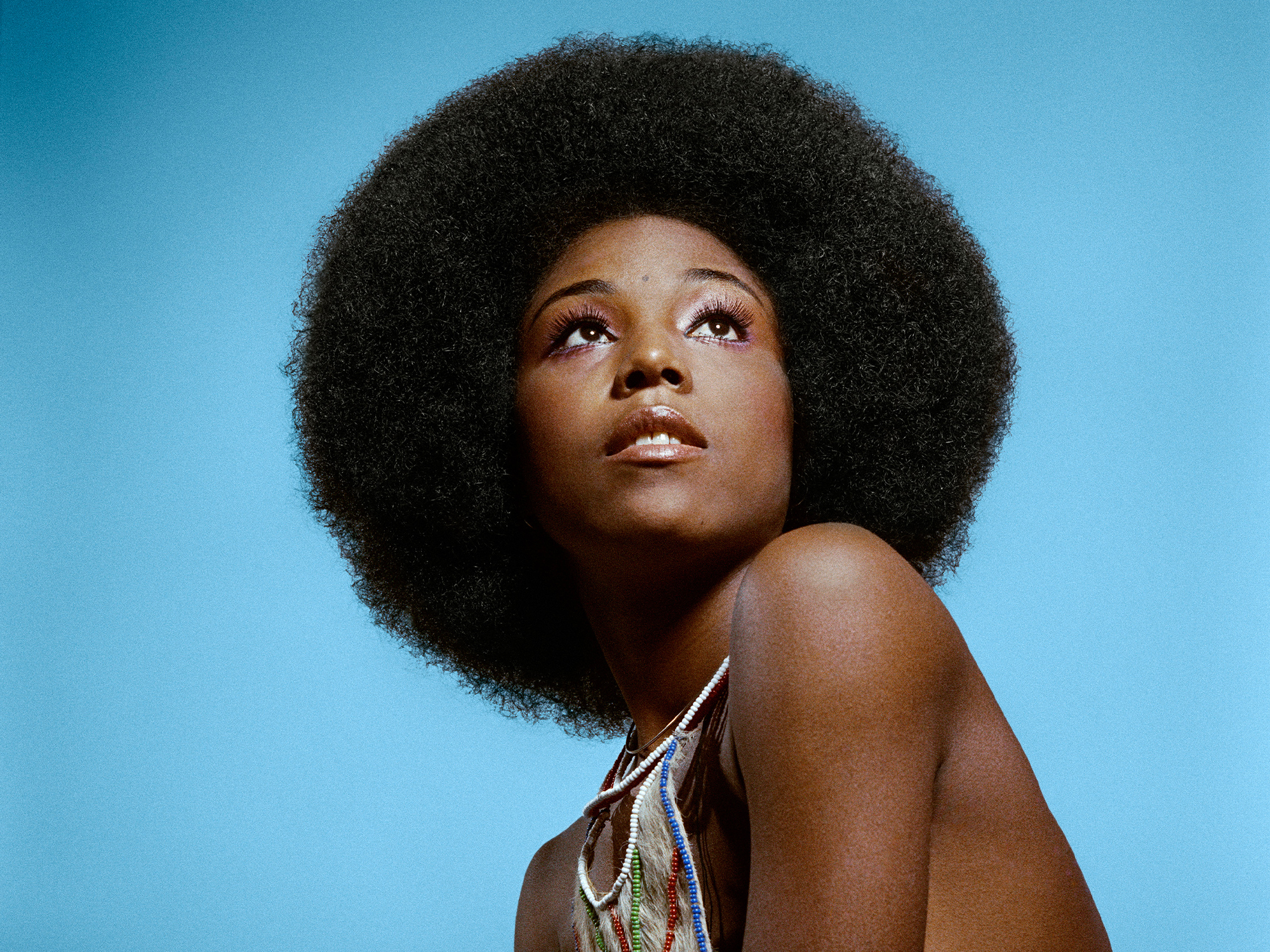 Woman afro hairstyle looking upward against blue background, bare shoulder visible.