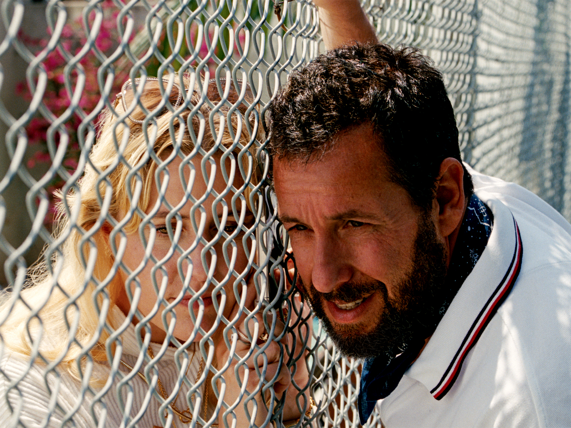 Man with beard in white polo shirt leaning against chain-link fence, smiling at camera in bright sunlight.