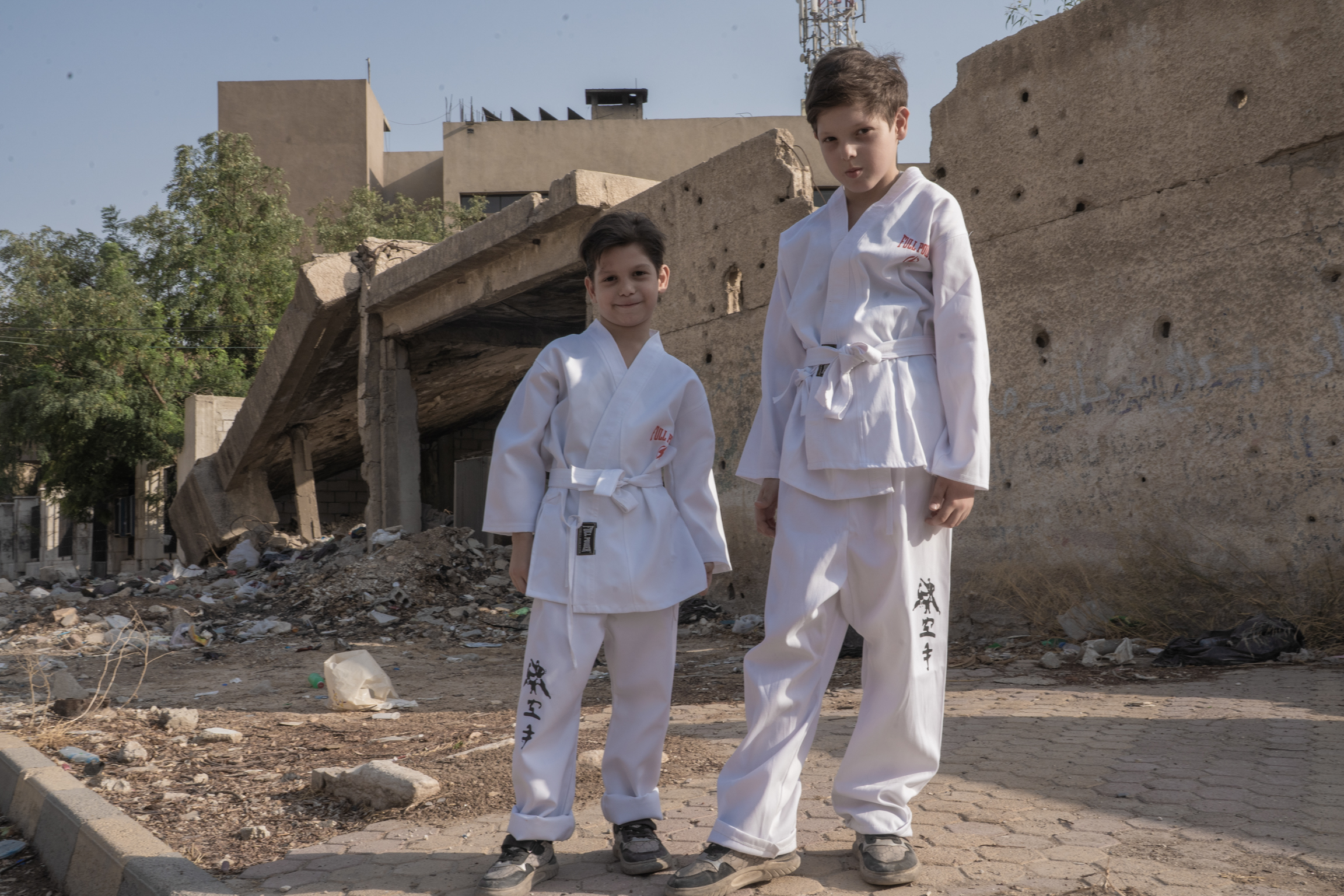 Two children in white martial arts uniforms standing on rubble-strewn ground with damaged concrete buildings in background.
