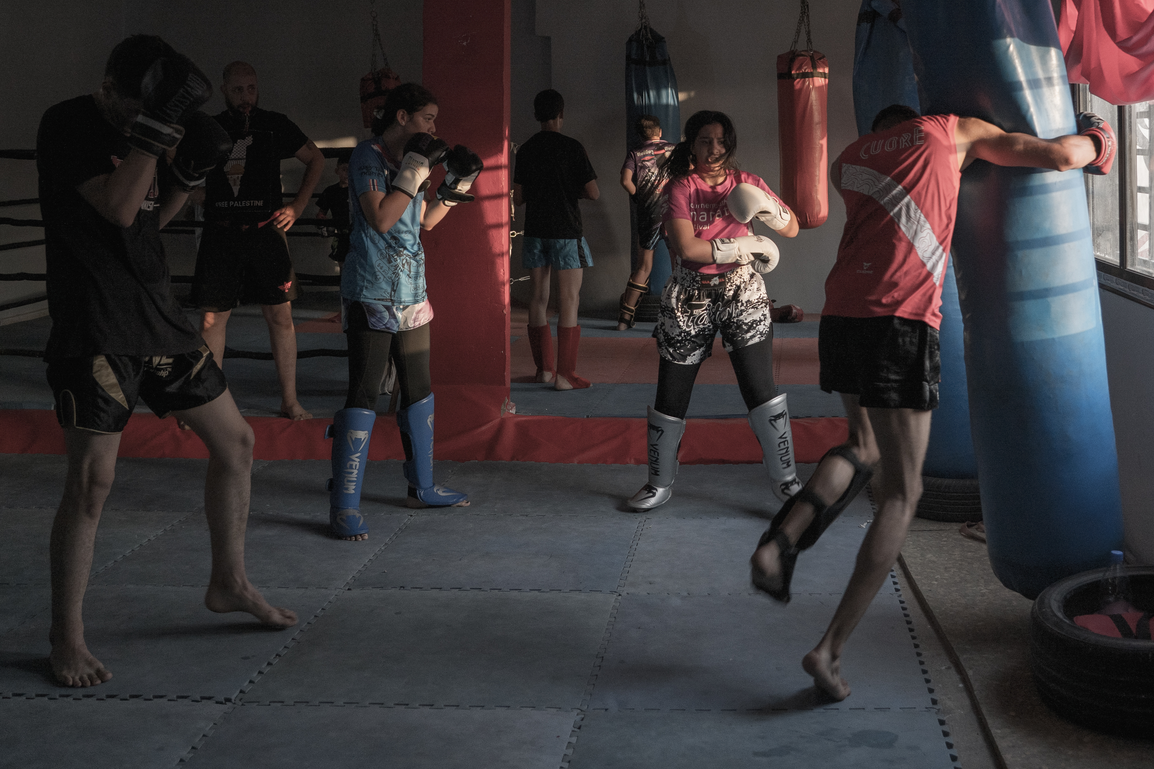 People training in boxing gym with red and blue heavy bags, dim lighting, concrete floor.
