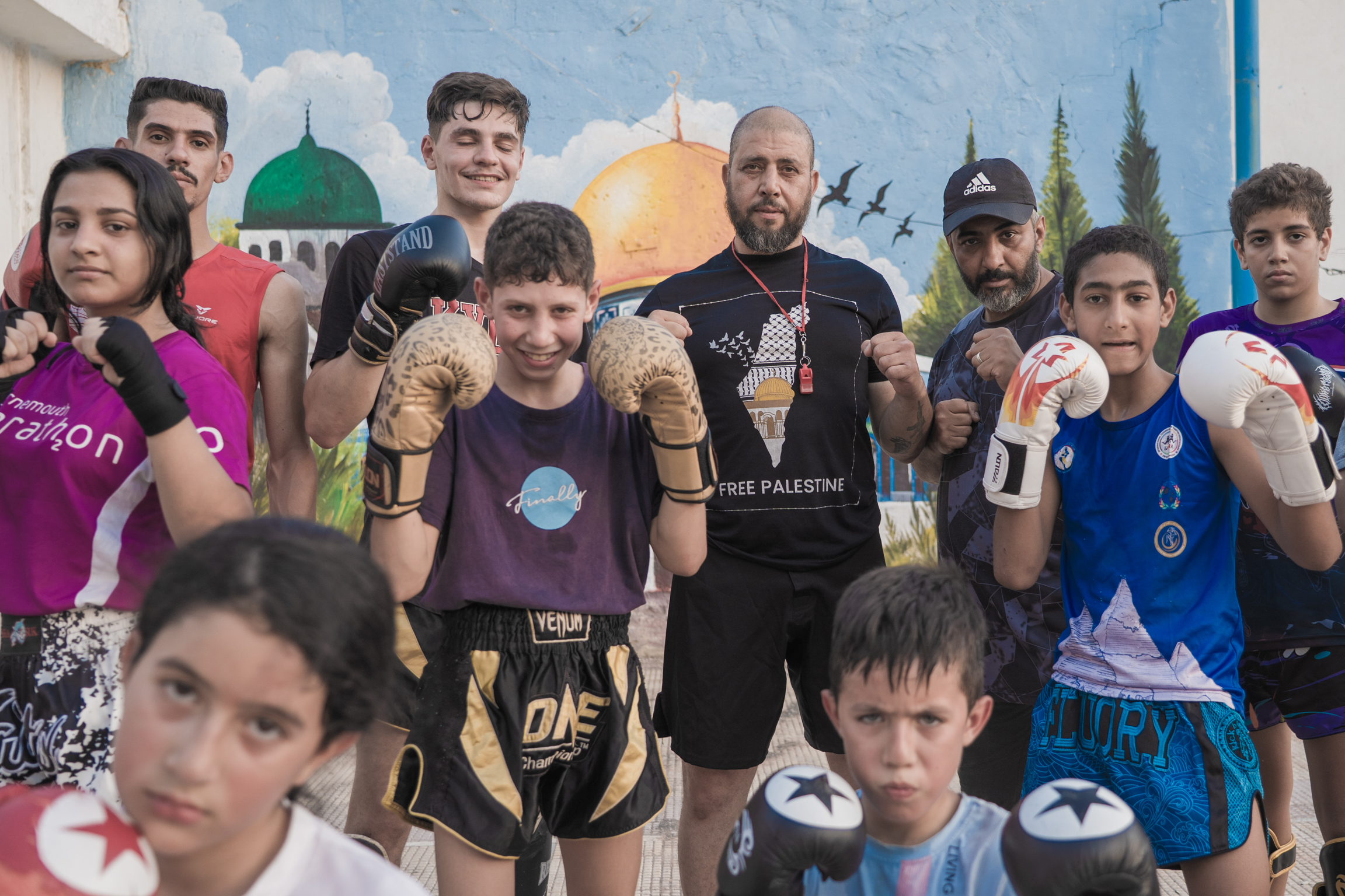 Group of children and adults in boxing gear posing together indoors, with colourful mural featuring domed buildings in background.