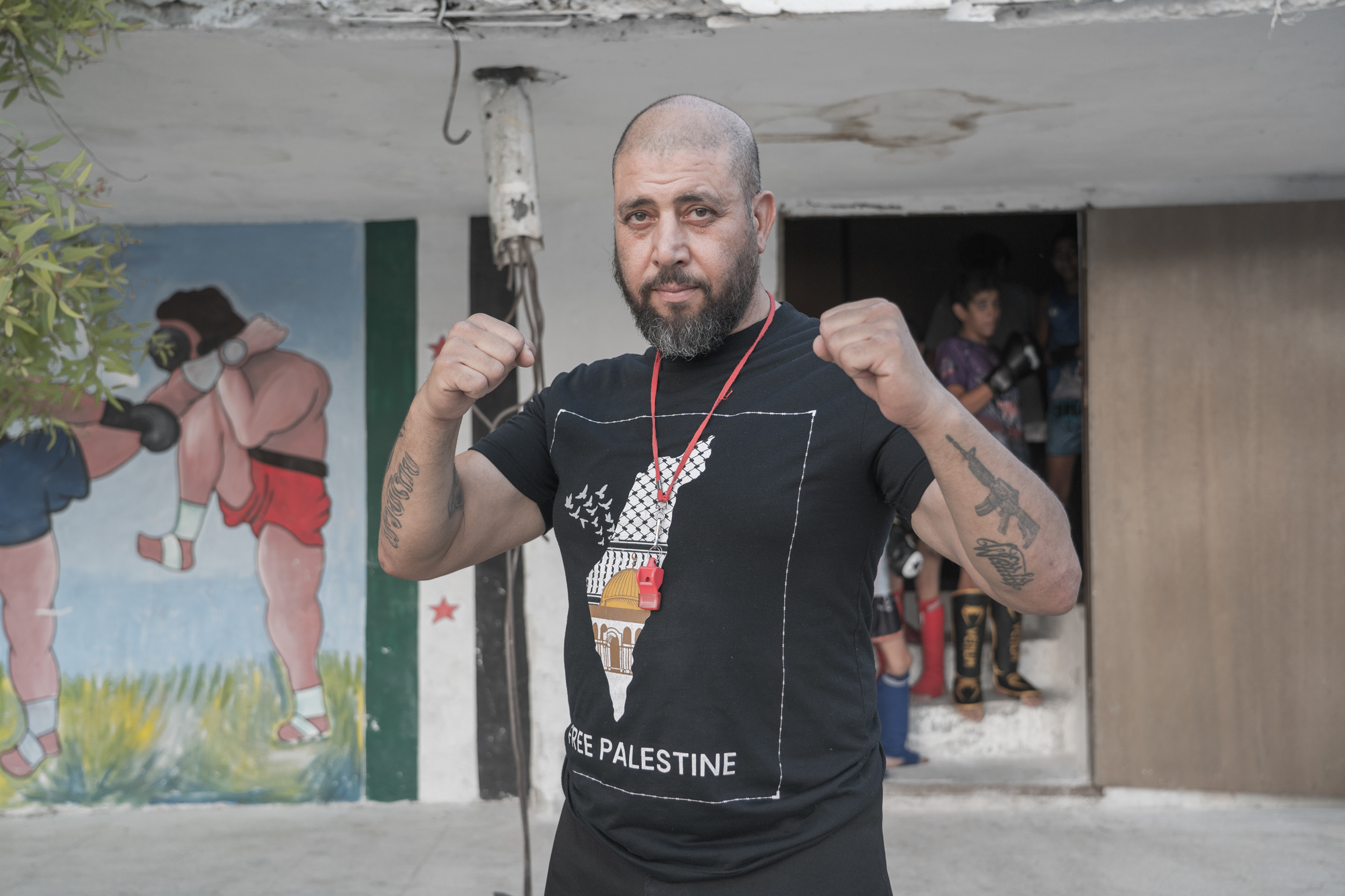 Bald bearded man in black Palestine t-shirt making fists, standing outside building with damaged ceiling and mural visible.