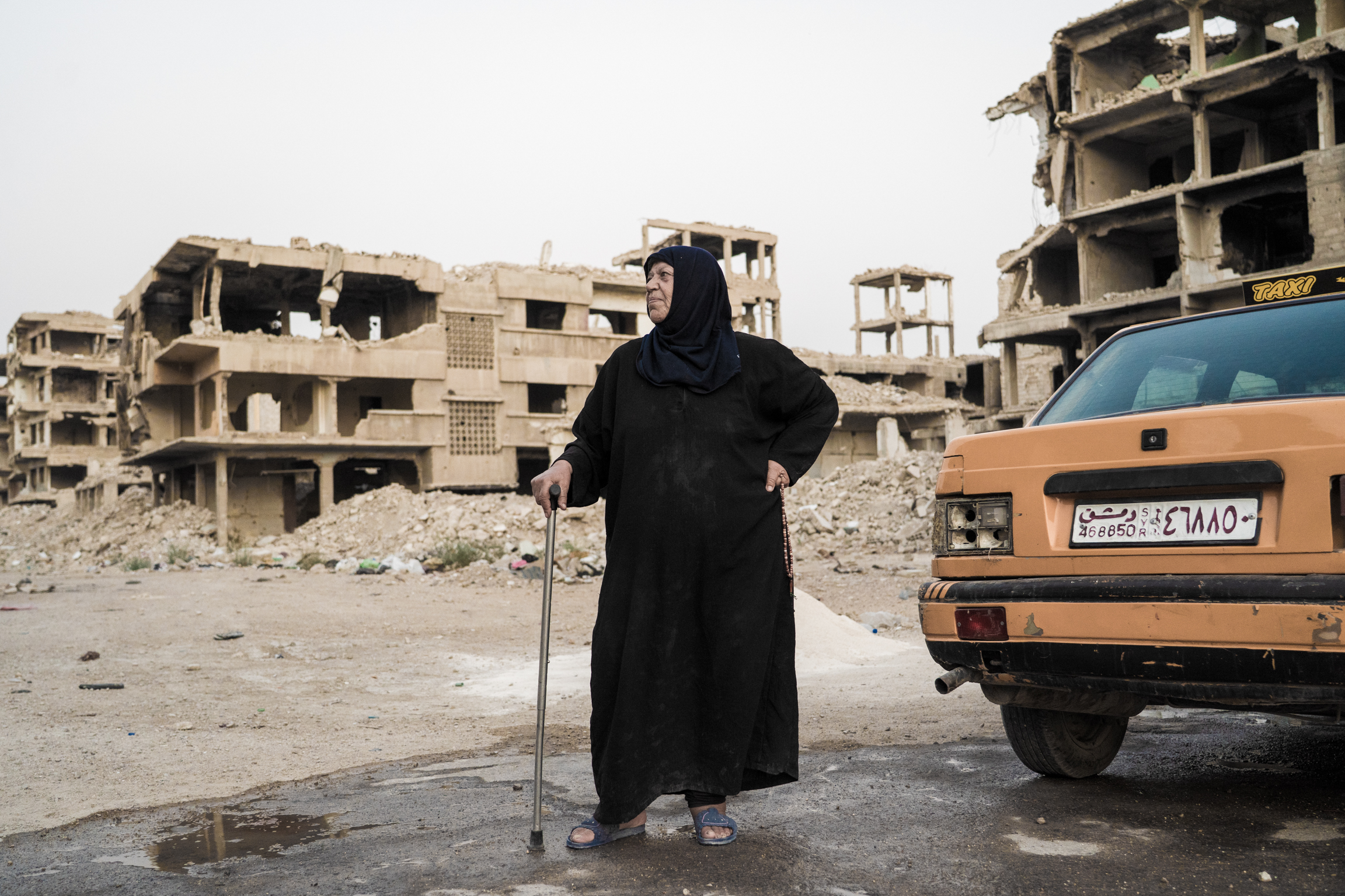 Person in black robes with walking stick stands near orange car amongst damaged concrete buildings and rubble.
