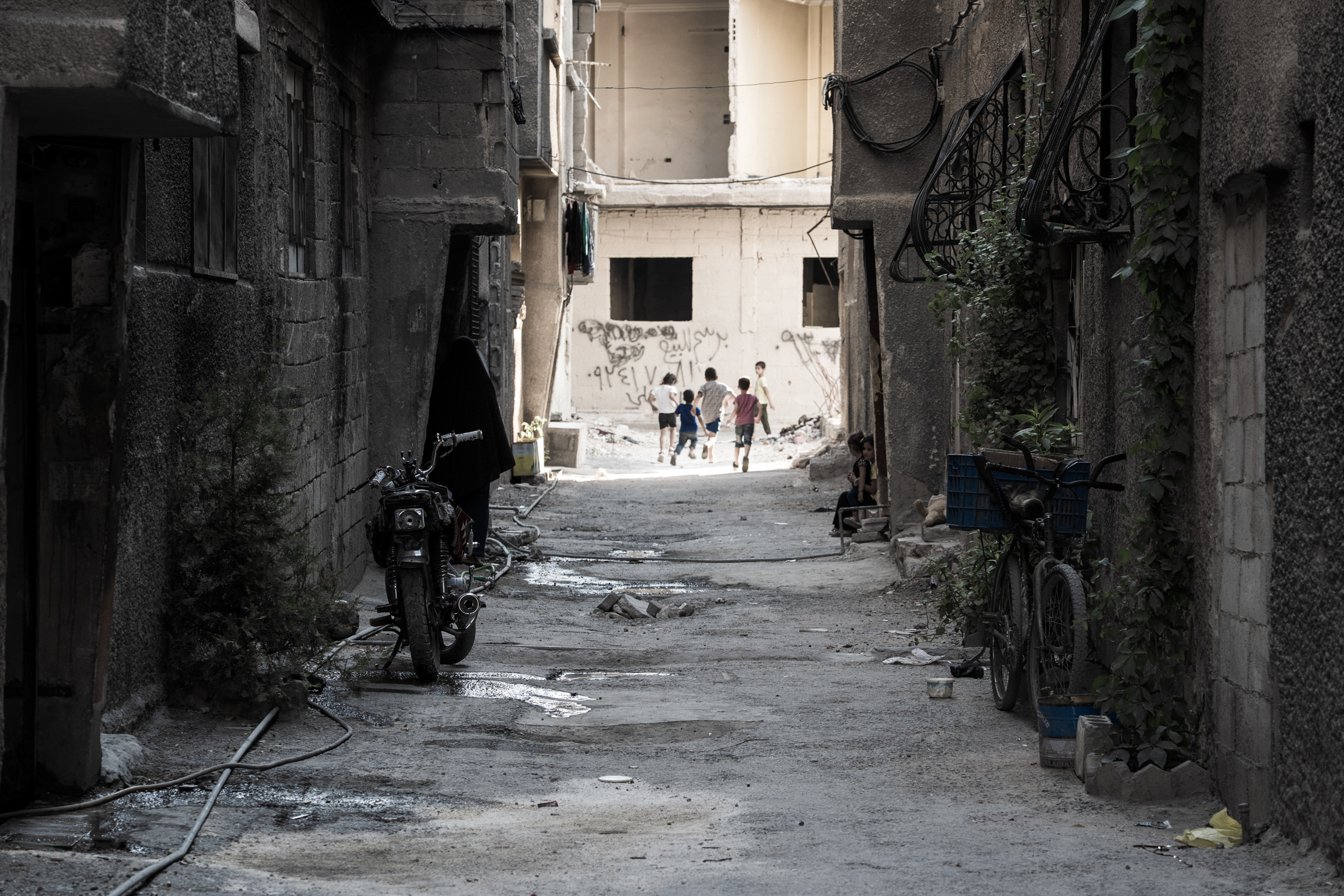 Narrow alley between damaged concrete buildings with debris scattered on ground. Two figures visible in bright courtyard at end of passage.
