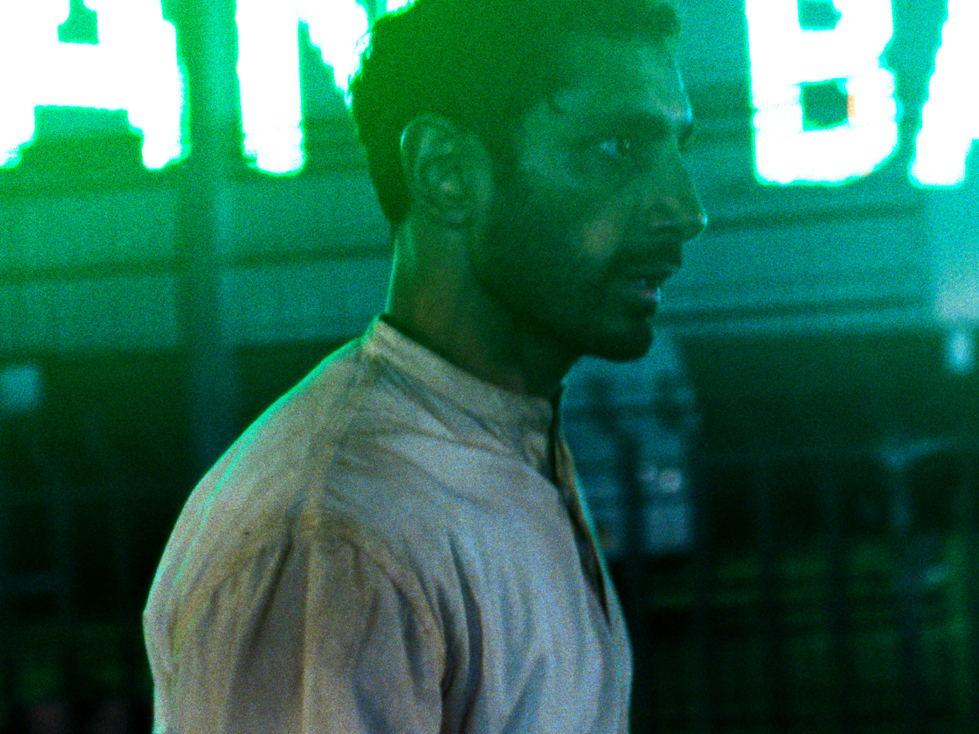 Man in white shirt with green and blue tinted lighting, blurred background with bright signage visible.