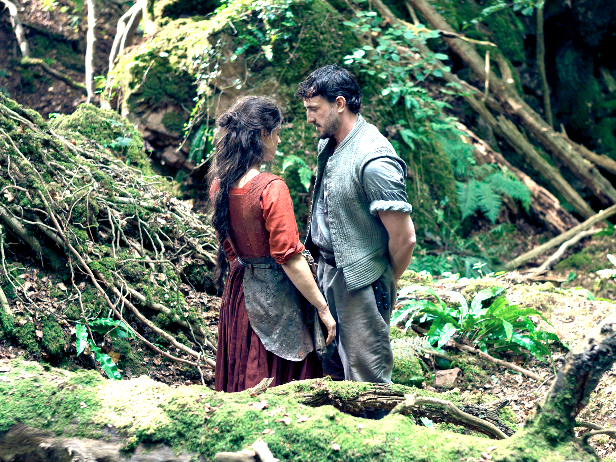 Woman in red dress and man in grey waistcoat standing close together in dense woodland with fallen logs and green moss.