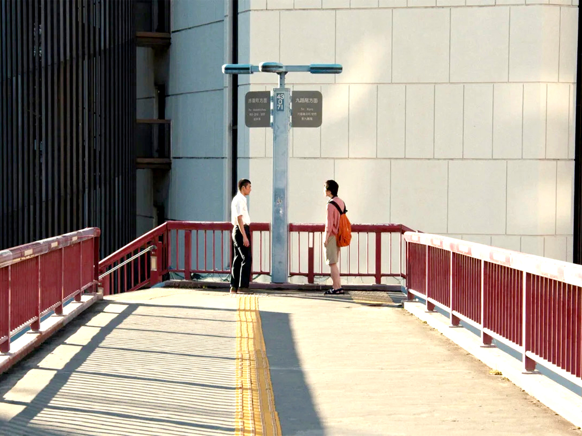 Two people standing on pedestrian bridge with red railings, white concrete surface, and grey building facade in background.