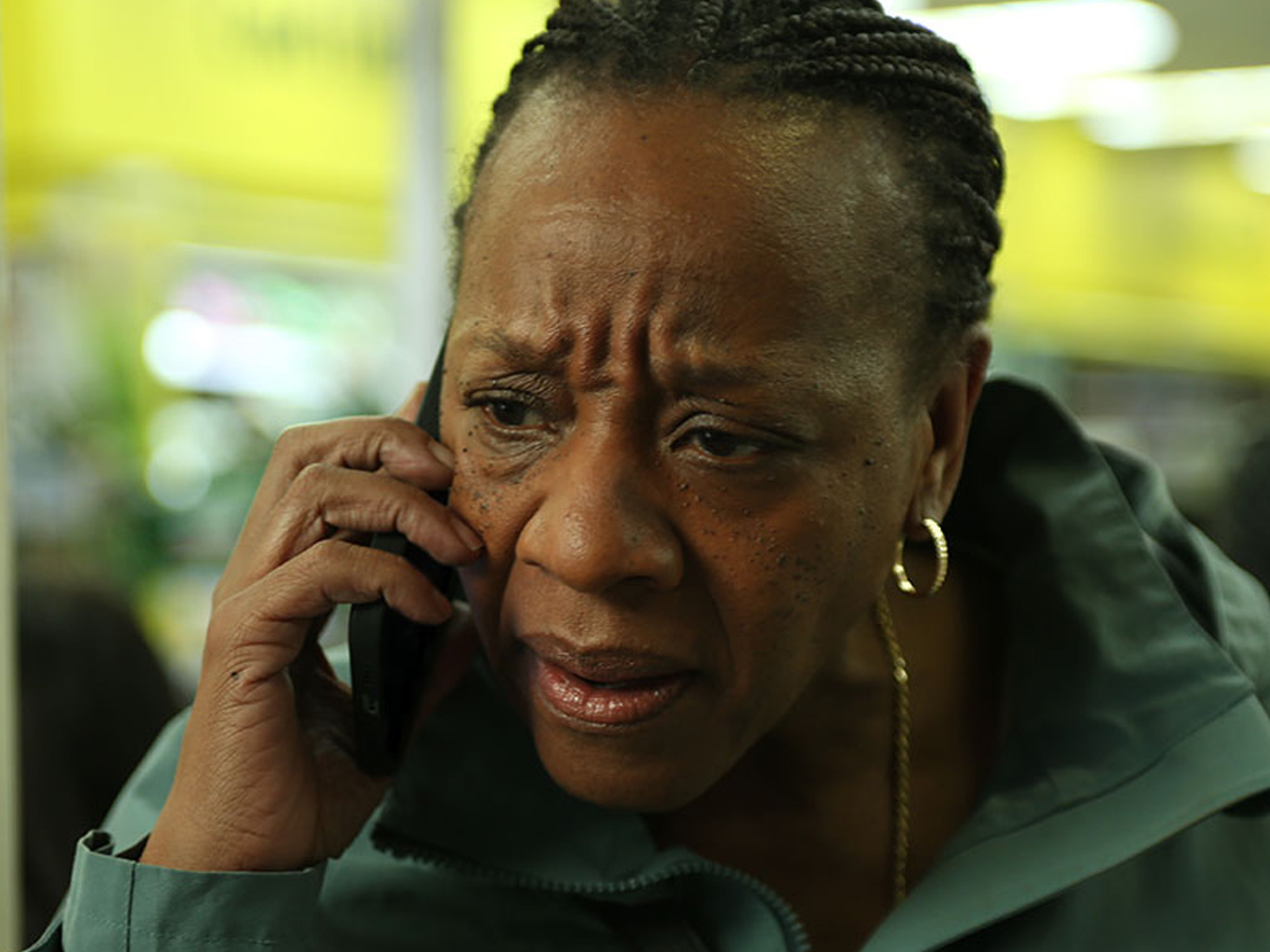 A serious-looking Black woman with braided hair, wearing a green jacket, speaking on a mobile phone.