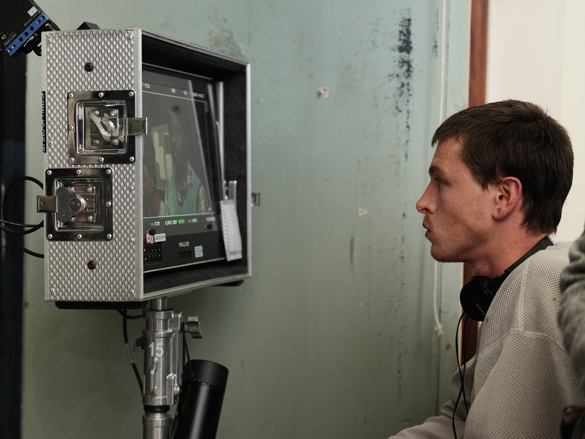 Man in light shirt operates industrial monitoring equipment with screen and controls mounted on wall in damaged room.