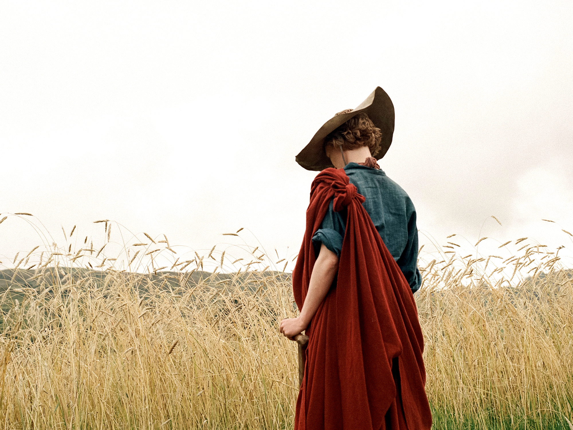 Person in dark hat, blue top, and red fabric wrap standing in golden grassland with birds flying overhead against pale sky.