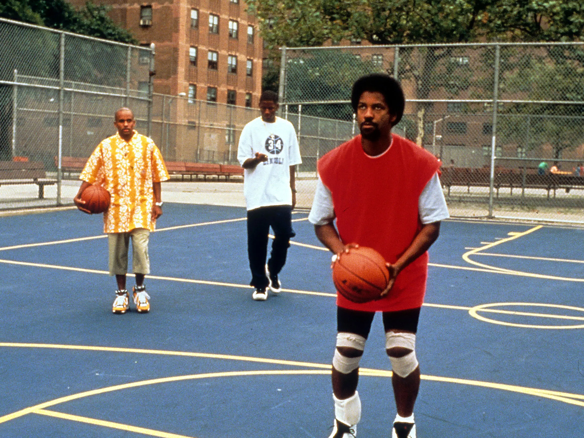 Denzel Washington wearing a red jersey holds a basketball on a blue basketball court. Two men stand behind him.