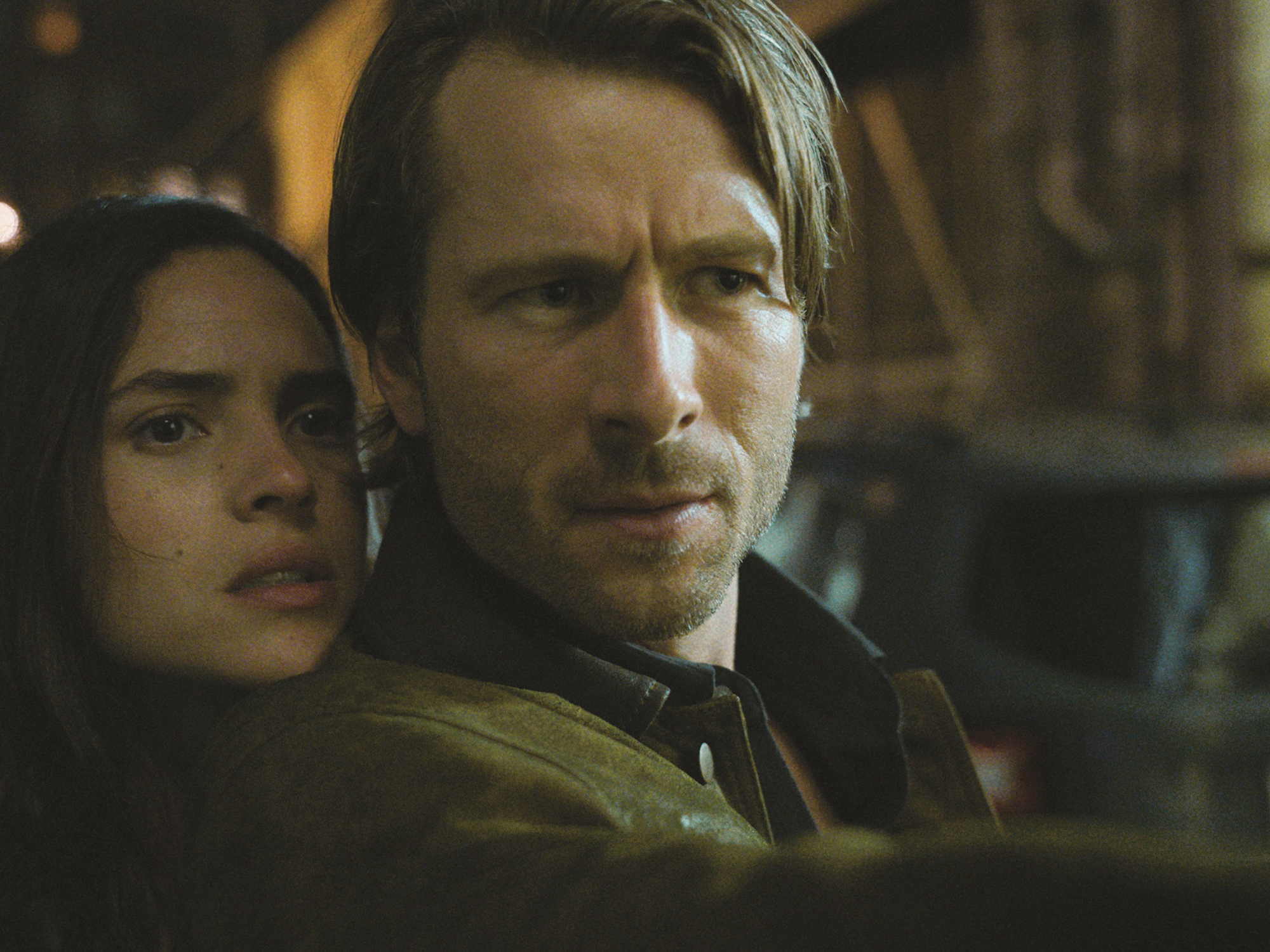 A man and woman in a dark, moody setting, the man has a serious expression.