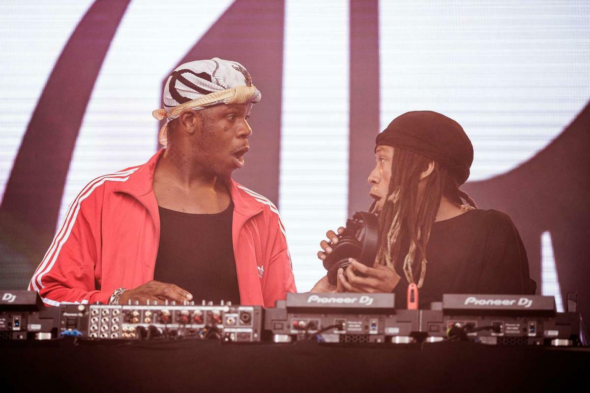 Two DJs performing at mixing deck, one in red jacket with head wrap, other in black with long hair and cap, large curved backdrop behind.