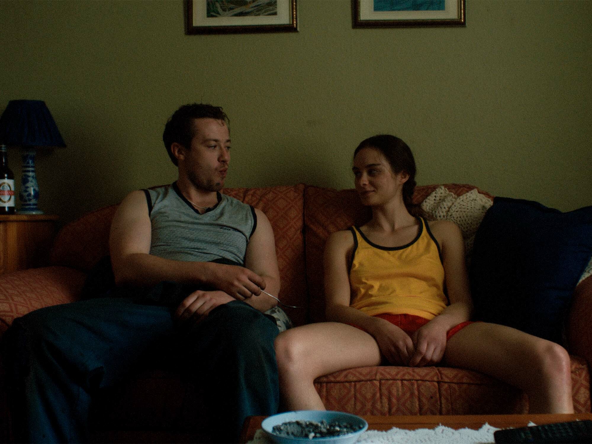 Two people sitting on a sofa, man wearing grey vest and woman wearing yellow top.