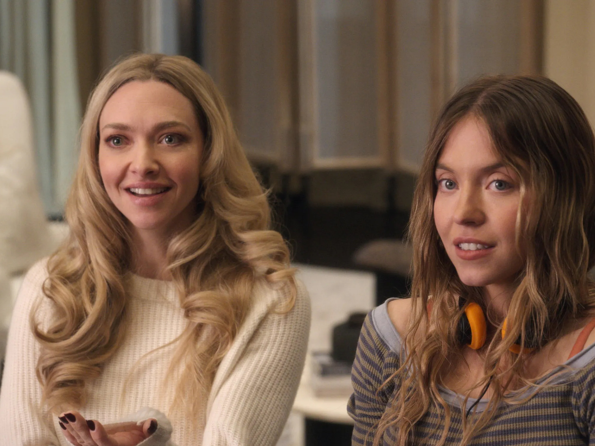 Amanda Seyfriend and Sydney Sweeney sitting together indoors - Amanda has blonde hair and is wearing a white jumper, Sydney has brunette hair and is wearing a grey striped top and orange headphones.