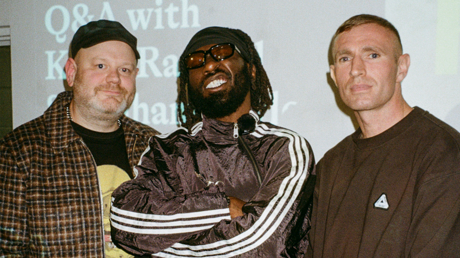 Three men posing together indoors - one in plaid jacket, one in dark tracksuit with sunglasses and dreadlocks, one in brown jumper.