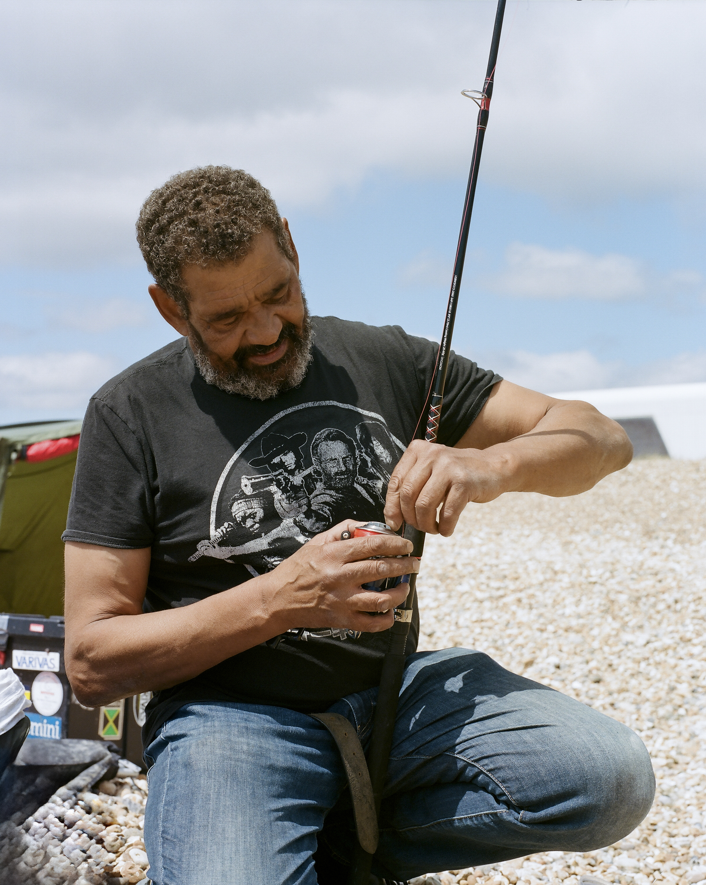 Bearded man in grey t-shirt handling small fish on pebble beach, fishing rod visible, cloudy sky background.