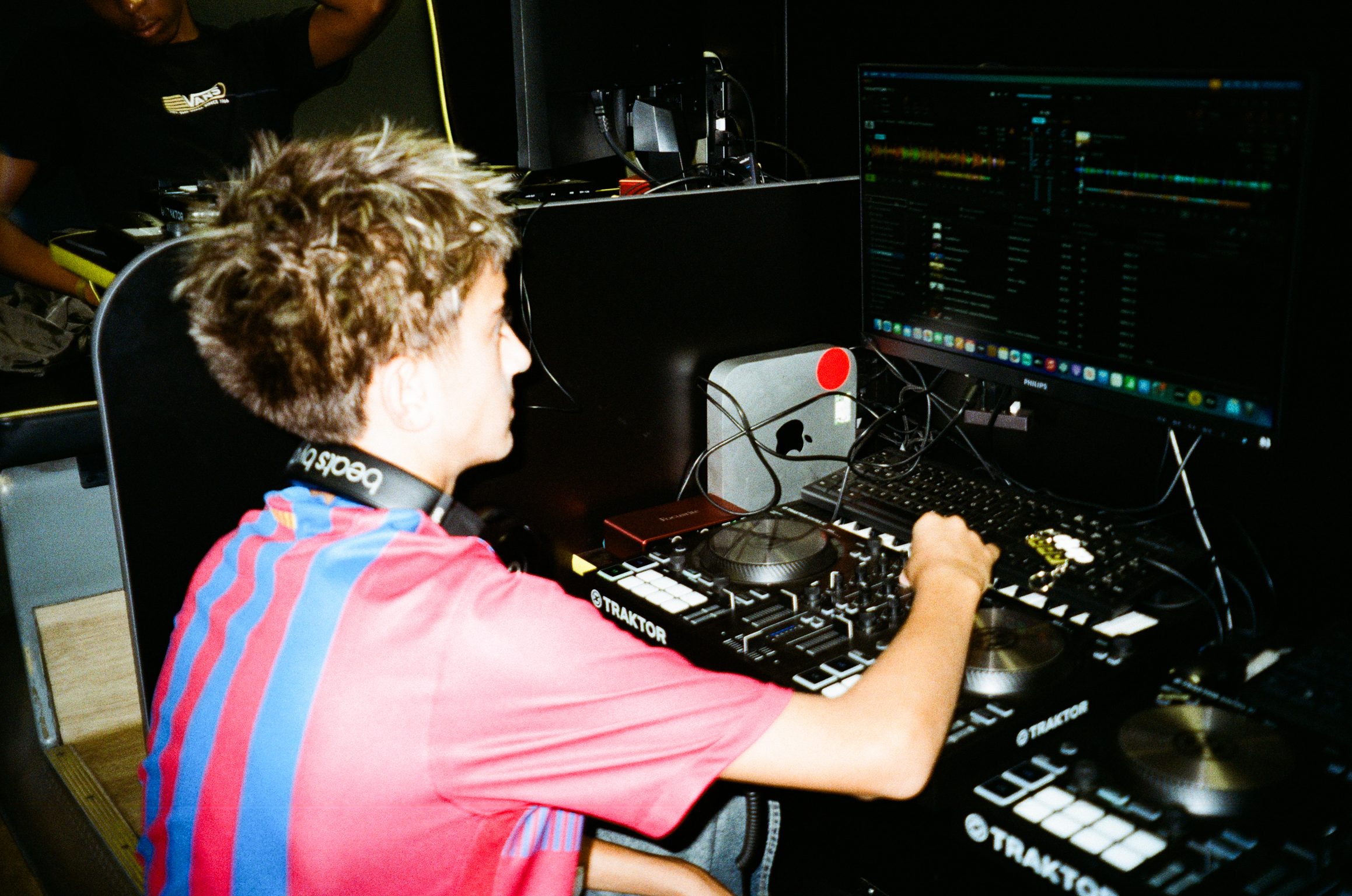 Person with curly blonde hair in pink and blue striped shirt operating DJ mixing desk with turntables in dimly lit venue.